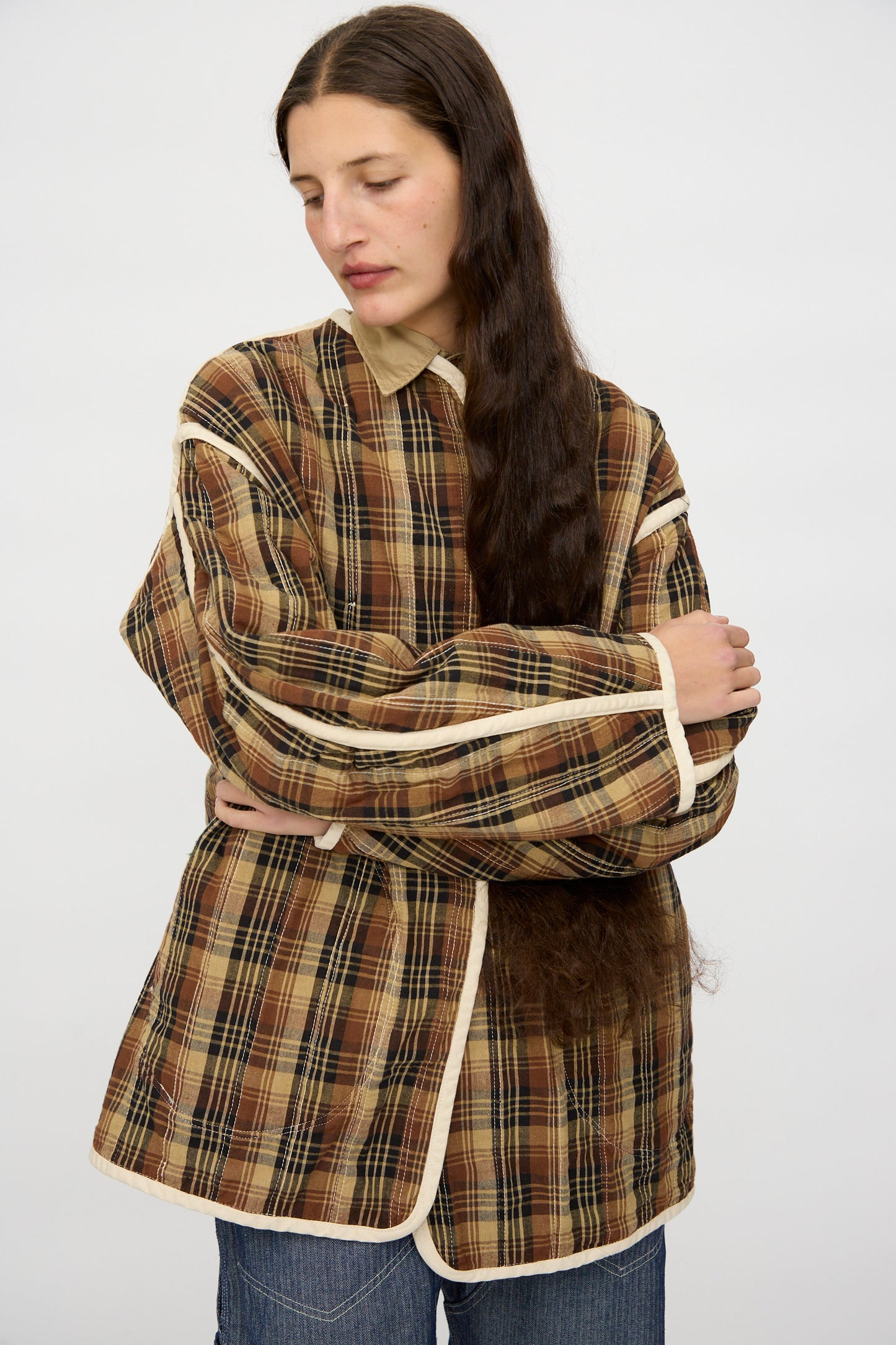 A person with long brown hair wears the As Ever Quilt Coat in HBT Natural, featuring an oversized fit and cotton batting, over a tan collared shirt, standing with arms crossed against a plain white background.