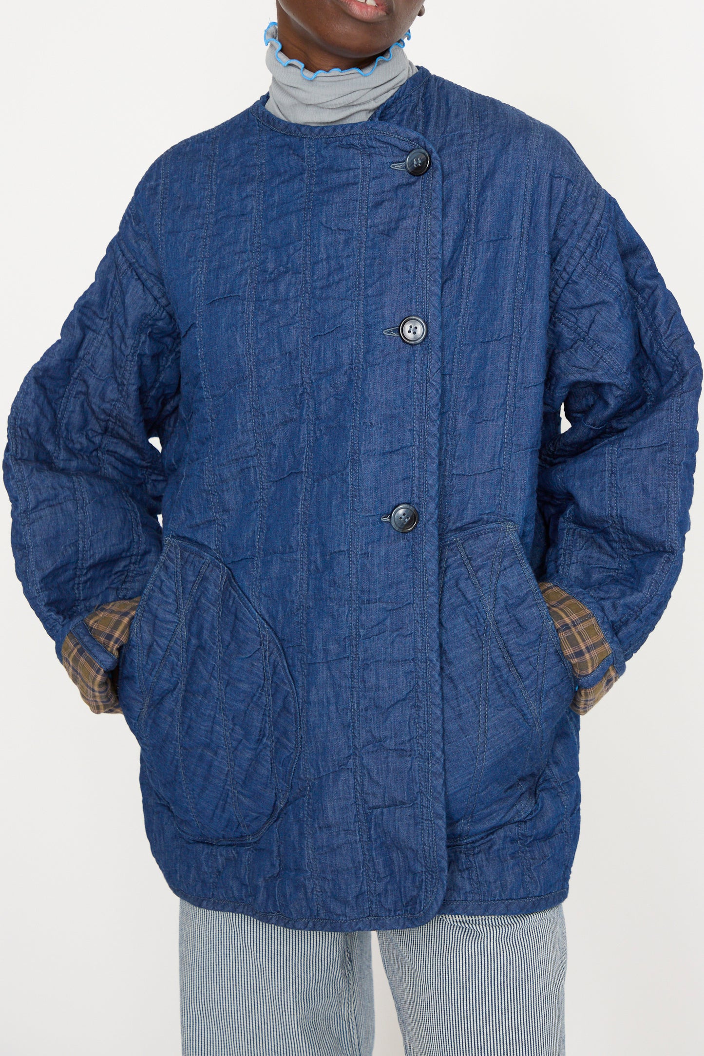 The individual wears an As Ever Quilt Jacket in Indigo / Plaid over a striped high-neck shirt, with their hands resting in the jacket's spacious pockets against a white backdrop. 