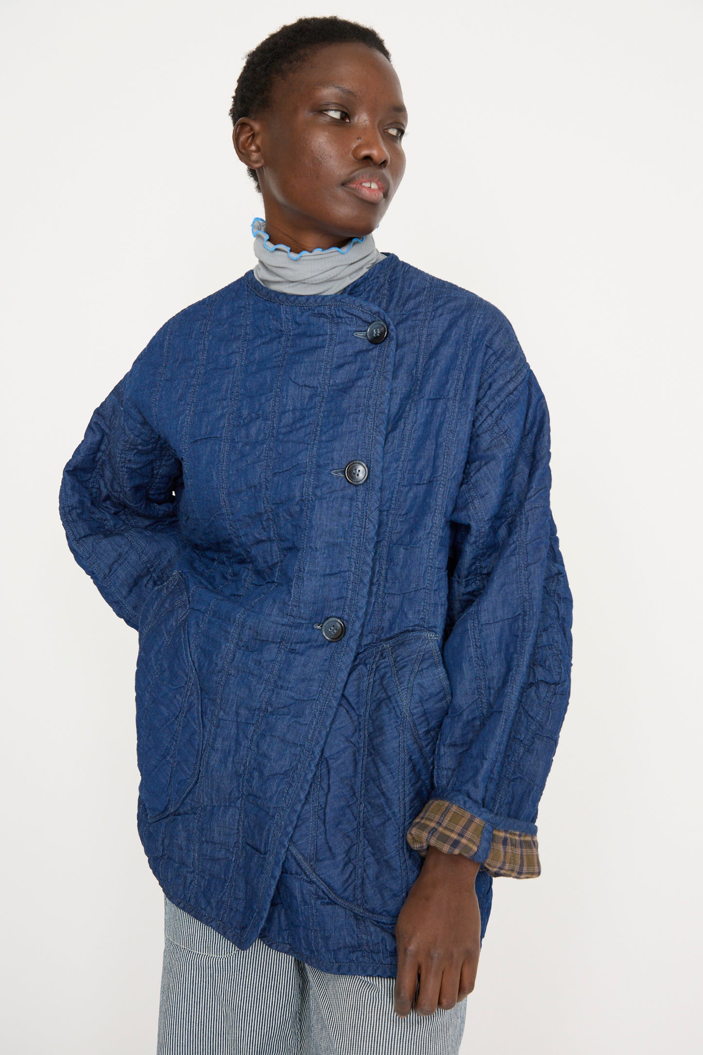 A person stands against a plain backdrop, wearing As Ever's Quilt Jacket in Indigo / Plaid, featuring asymmetrical buttons, looking to the side. 