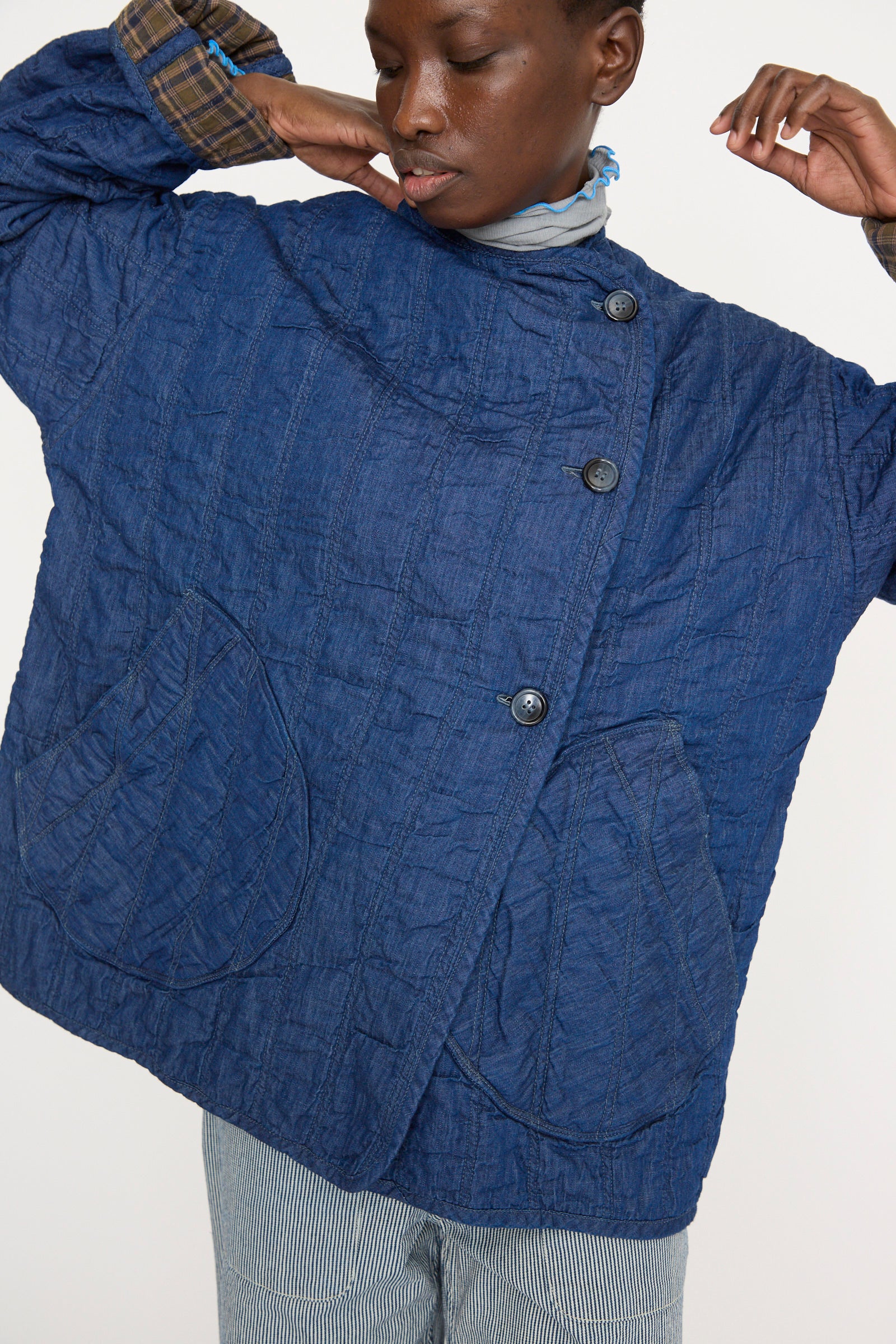 The model is wearing the As Ever Quilt Jacket in Indigo / Plaid, featuring large pockets and a button-up front. The plaid lining at the cuffs shows its reversible cotton design.