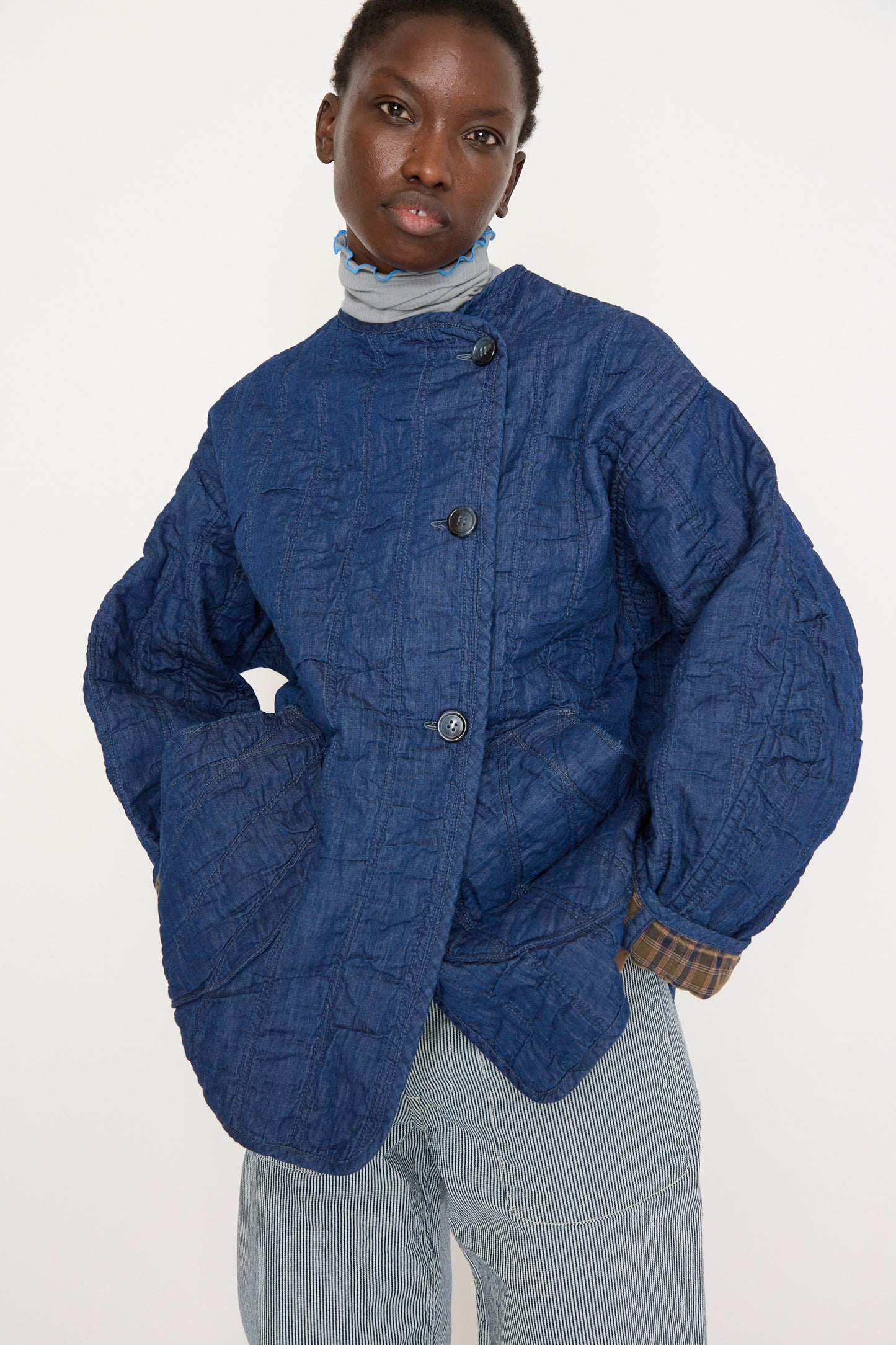 A person poses with hands in pockets, wearing As Ever's Quilt Jacket in Indigo / Plaid, featuring a textured quilted design with large buttons. The jacket is paired with a striped turtleneck and gray striped pants.