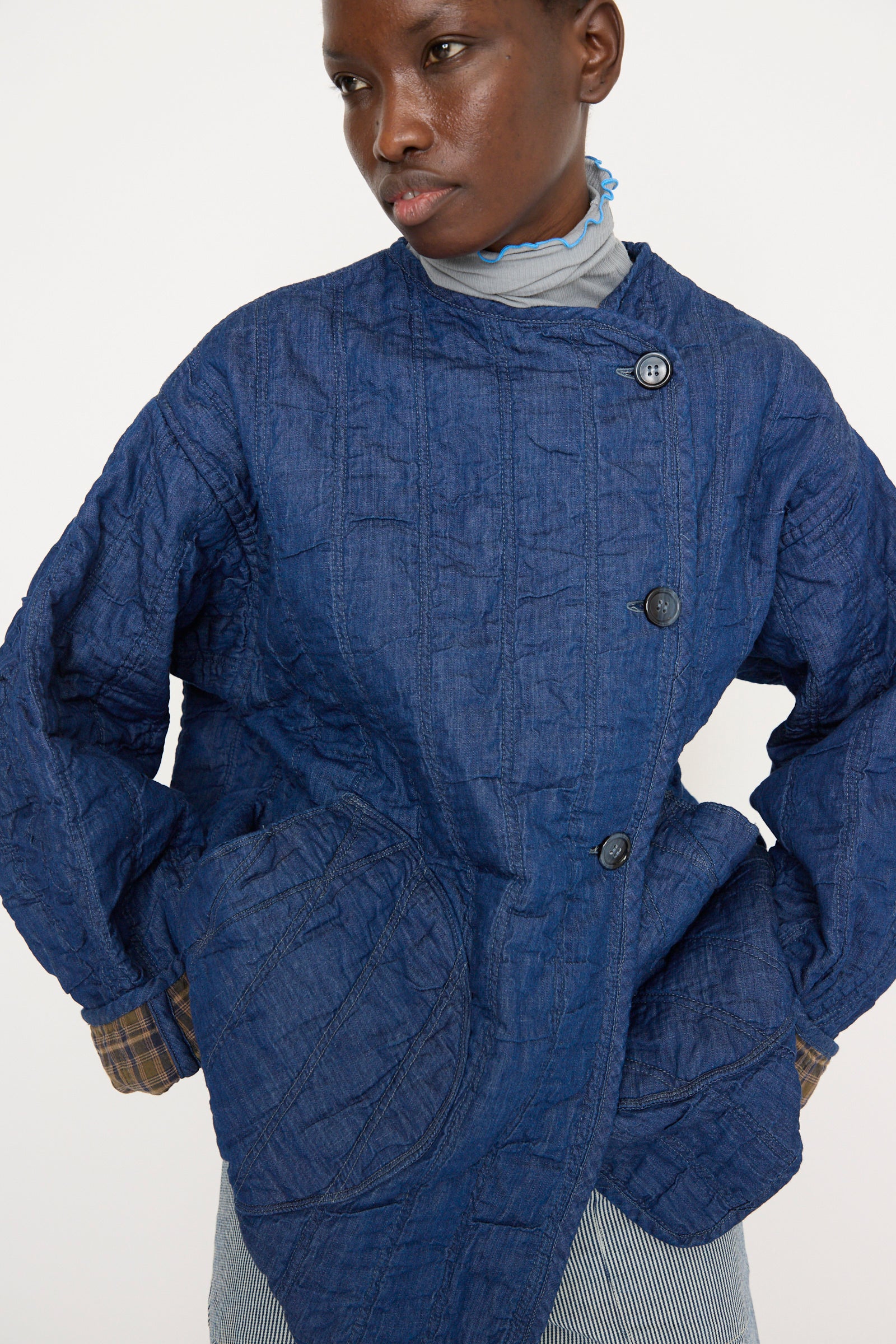 A person is wearing the As Ever Quilt Jacket in Indigo/Plaid, which features a textured cotton material, reversible design with buttons and plaid cuff details.