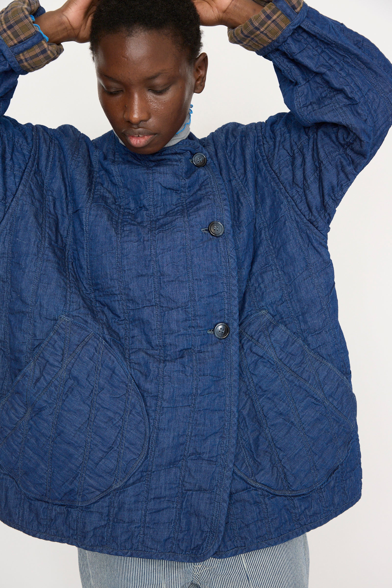 A person wears a blue As Ever Quilt Jacket in Indigo with large pockets and buttons, paired with striped pants, against a plain background.