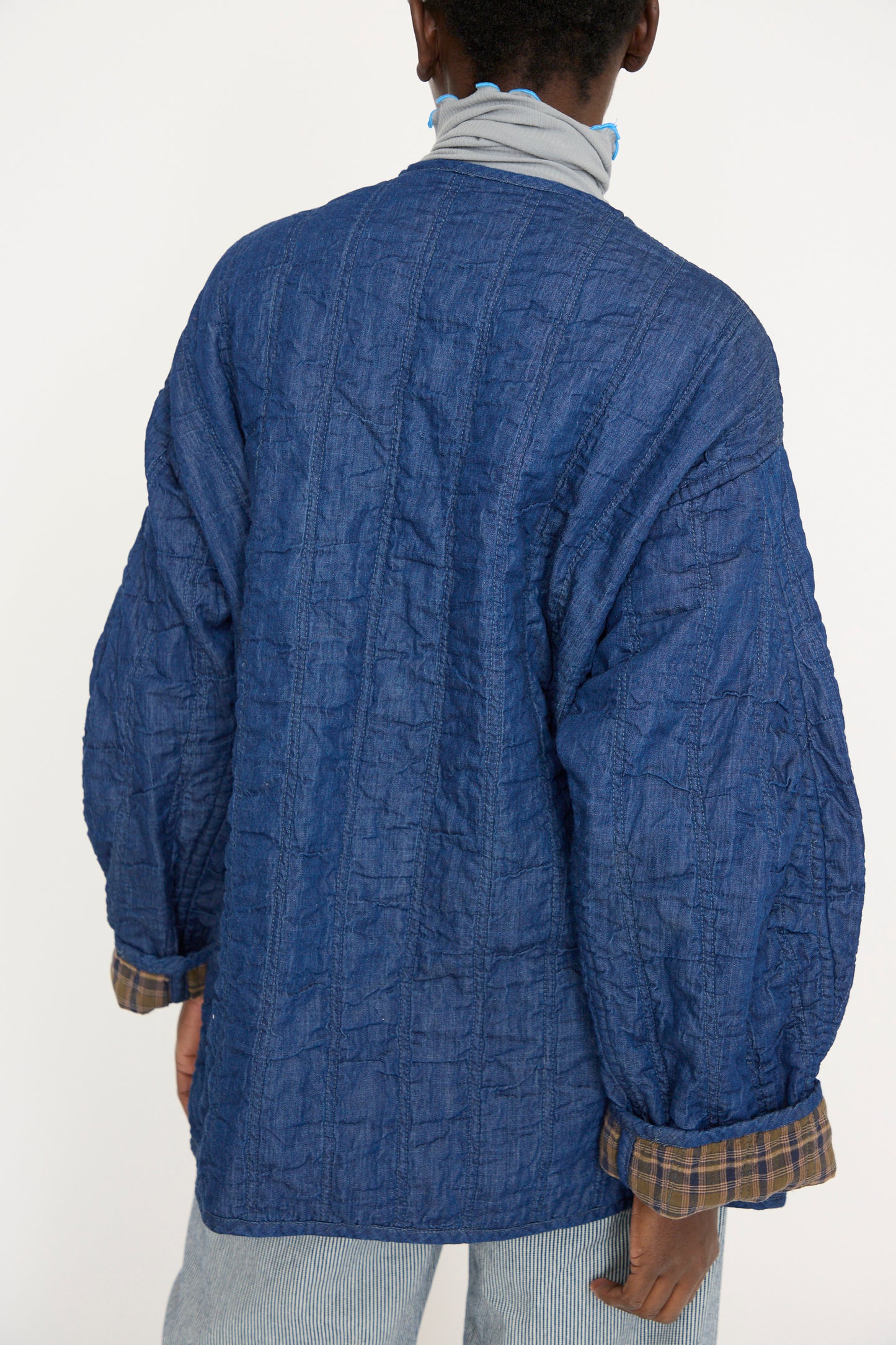 A person wears the As Ever Quilt Jacket in Indigo / Plaid, featuring a textured, oversized design with rolled-up plaid cuffs, facing away against a plain background. 