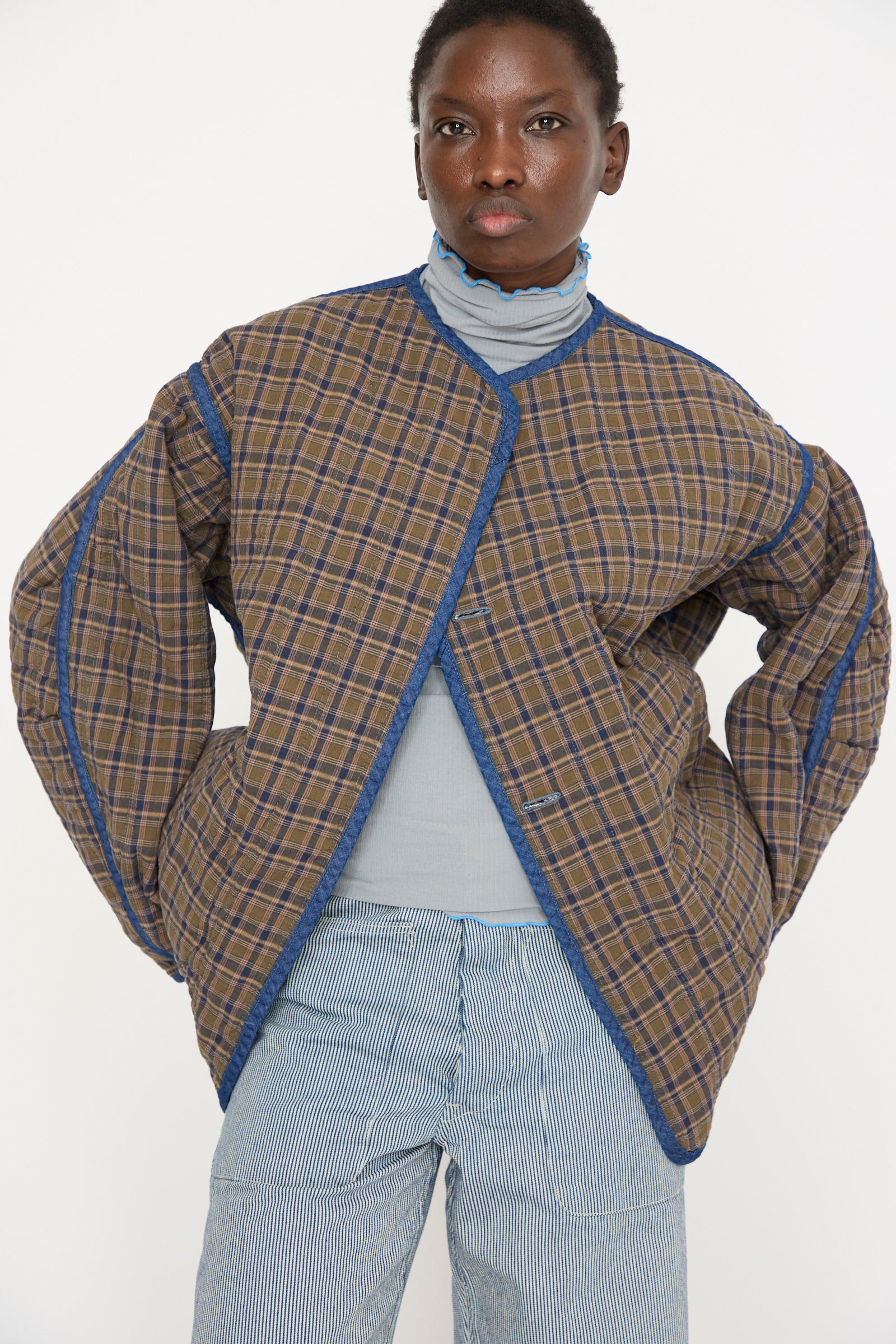 A person stands against a plain background wearing As Ever's Quilt Jacket in Indigo/Plaid over a light blue top and striped pants.