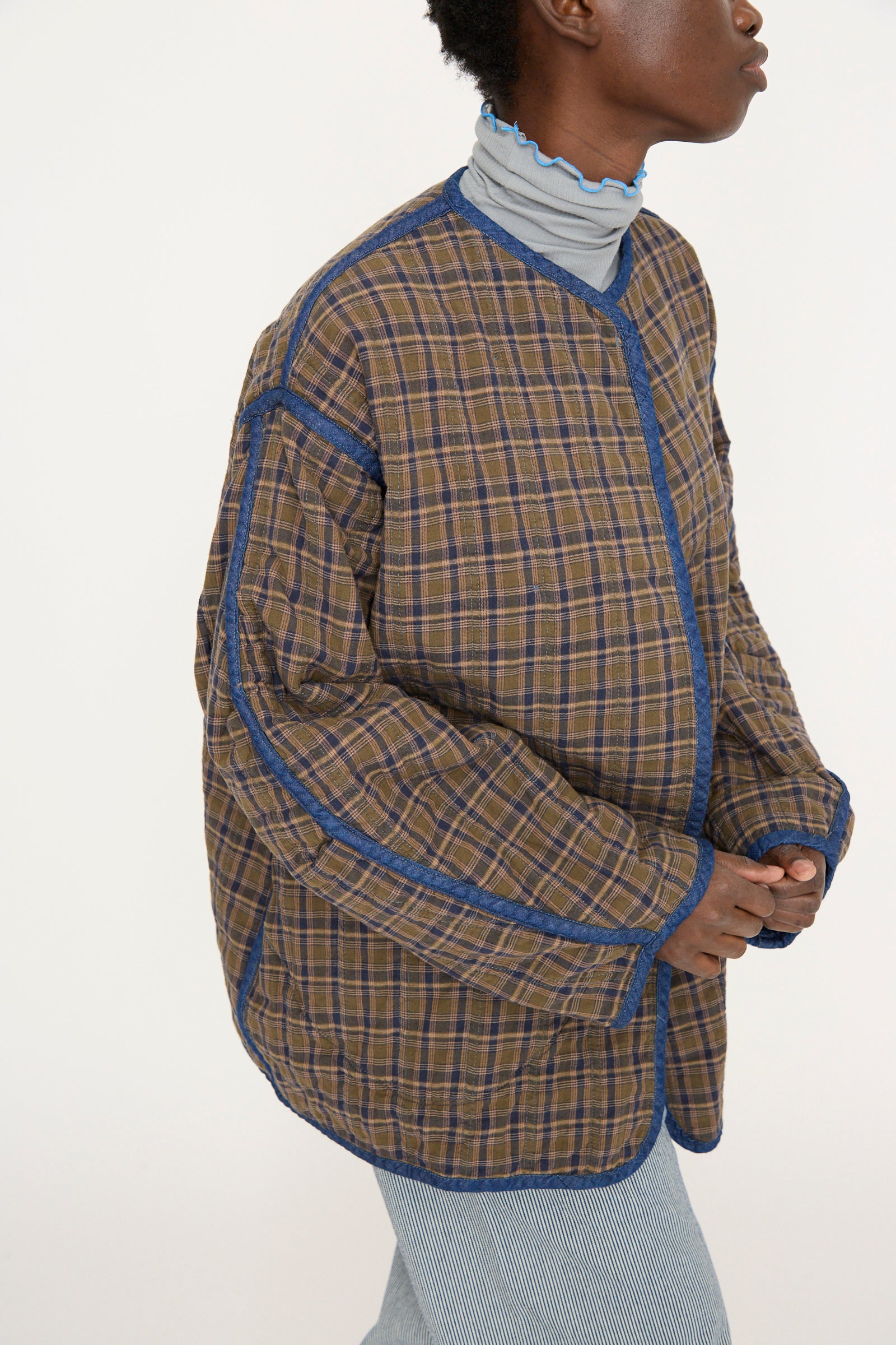 A person in the Quilt Jacket in Indigo / Plaid by As Ever, reserved to the brown plaid side with blue trim, layered over a light blue turtleneck, stands in a side profile pose.