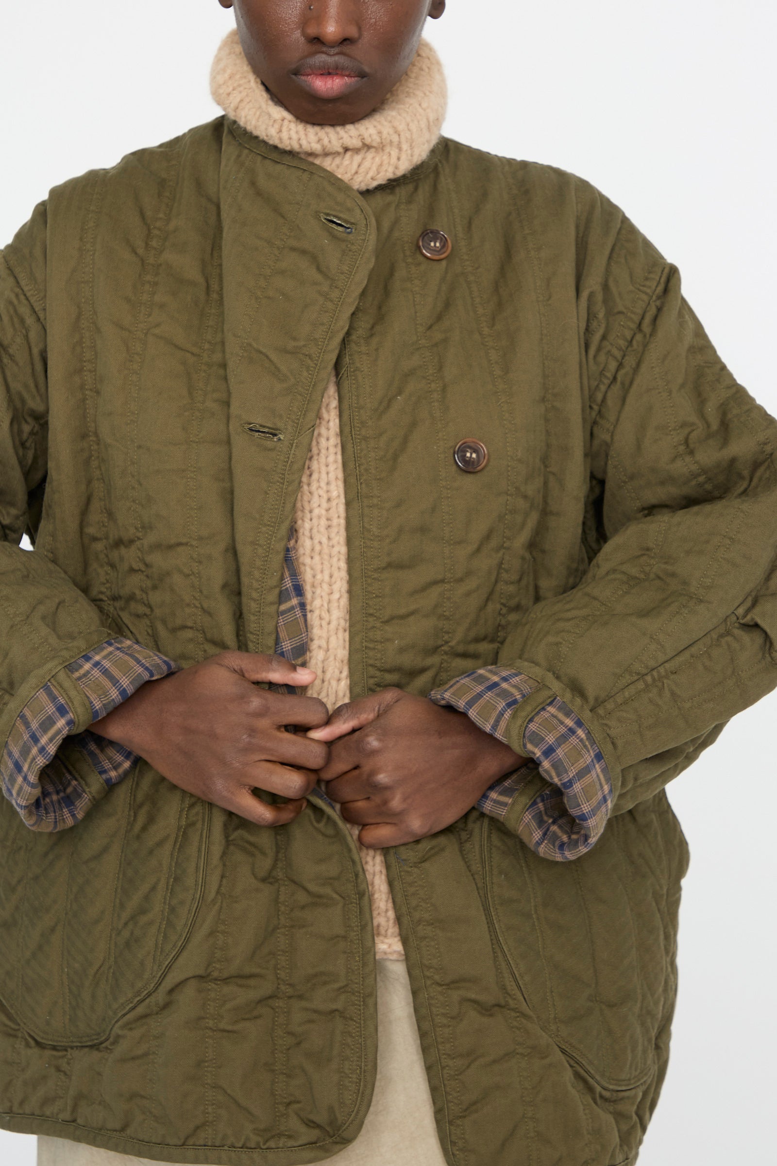 A person adjusts the button of an oversized Quilt Jacket in Olive by As Ever, worn over a beige knit sweater.