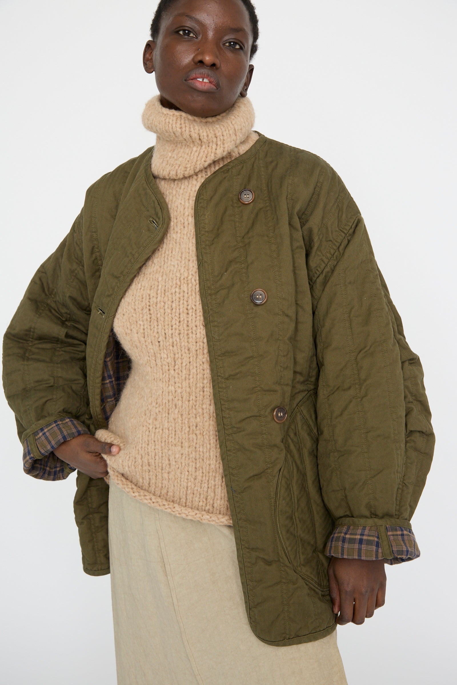 Against a plain background, a person is wearing As Ever's Quilt Jacket in Olive over a beige turtleneck sweater and matching skirt.