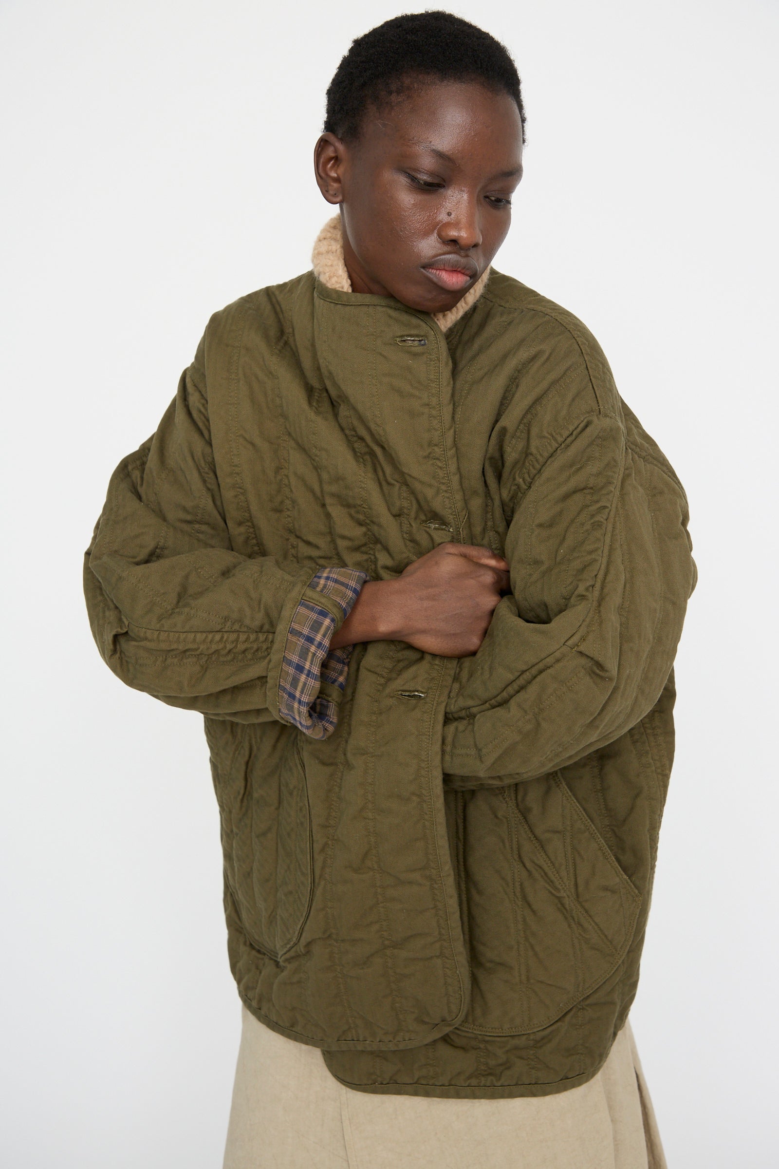 A person stands against a plain background wearing the As Ever Quilt Jacket in Olive, an oversized green quilted jacket with plaid lining.