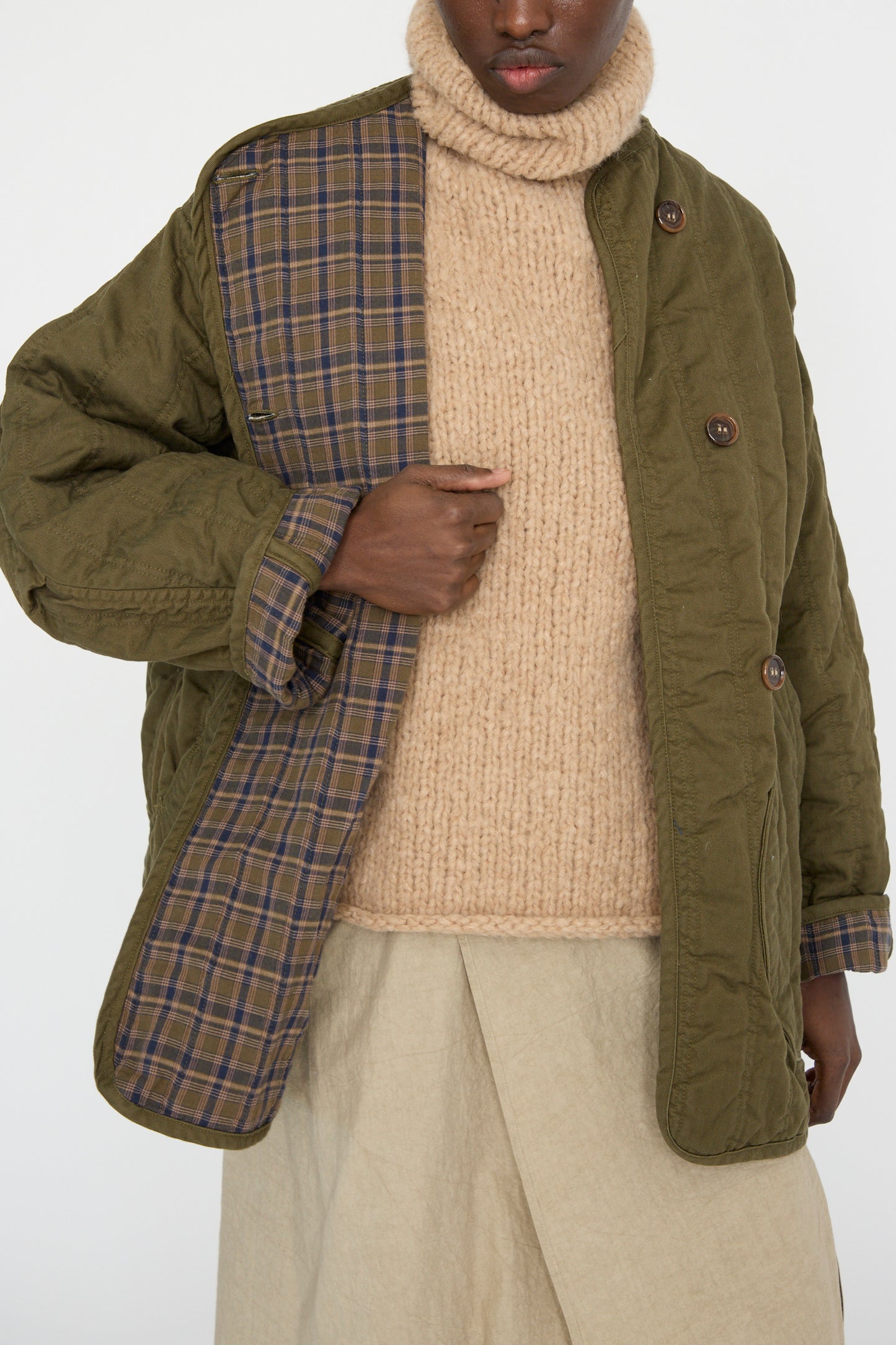 A person shows the plaid lining of an oversized Quilt Jacket in Olive by As Ever, wearing a tan knit turtleneck and beige pants.