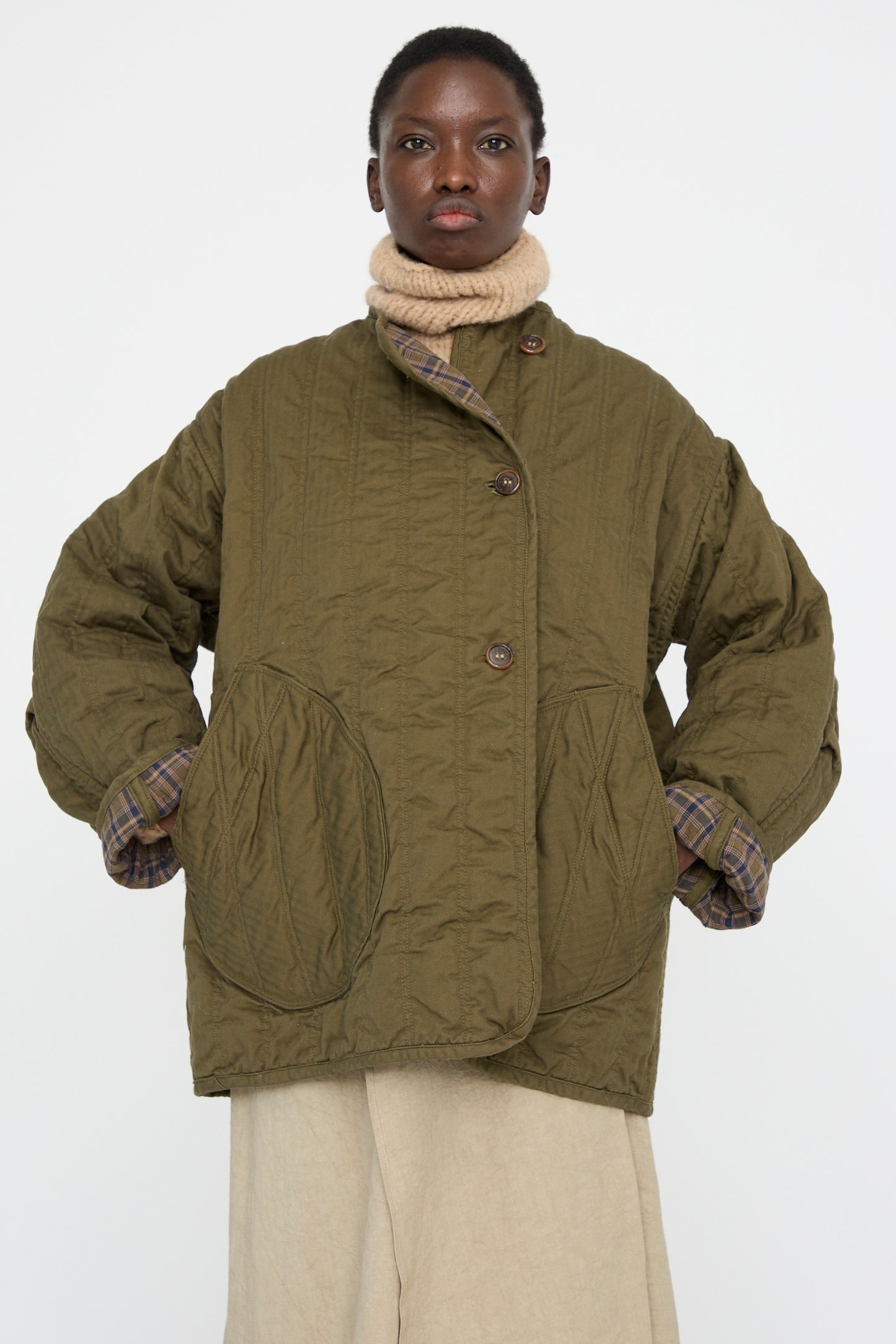 A model wearing the As Ever Quilt Jacket in Olive, a reversible vintage cotton piece with large pockets, over a beige turtleneck and plaid sleeves, set against a plain background.