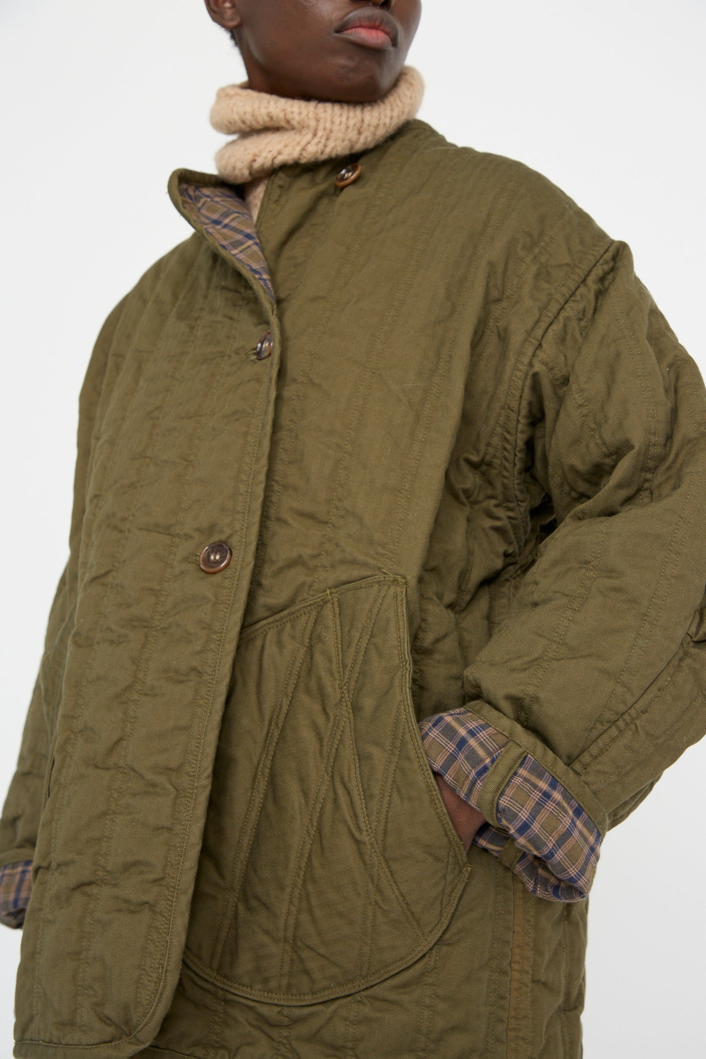 A person models the As Ever Quilt Jacket in Olive, with one hand in its large front pocket, revealing the plaid inner lining of this reversible garment.