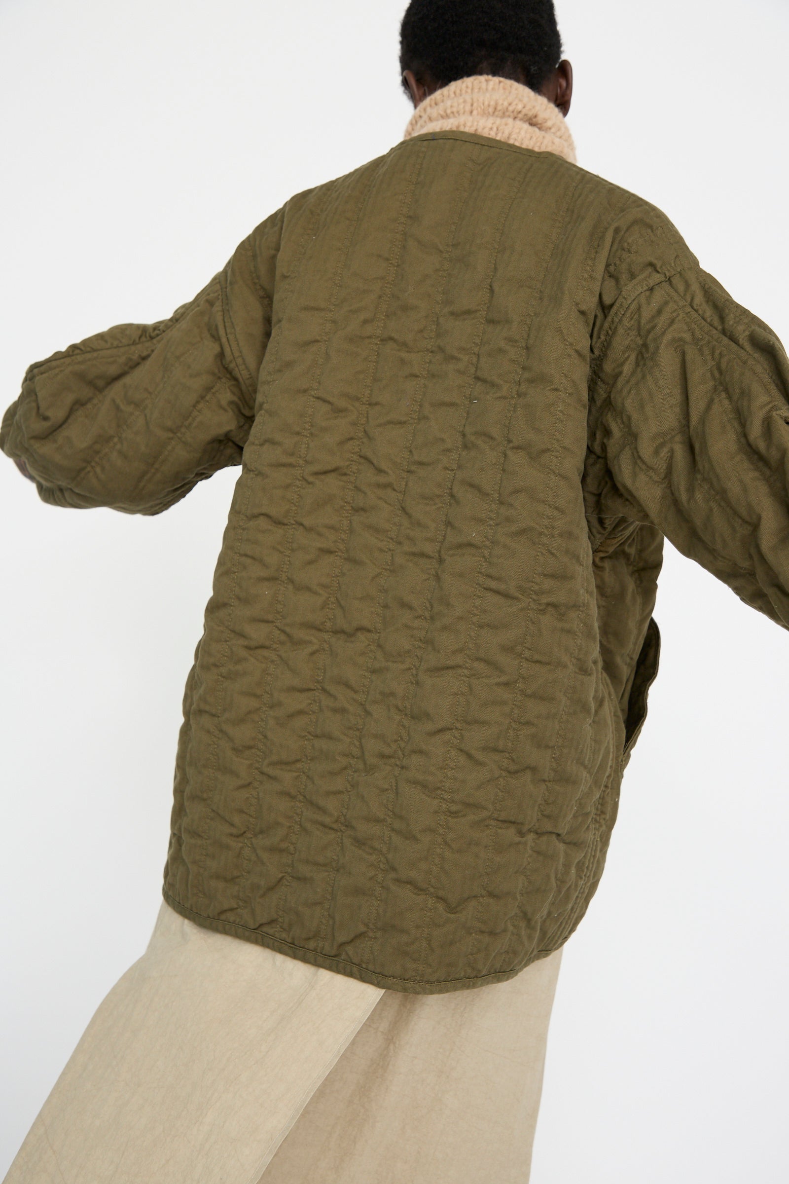 A person is seen from behind wearing the As Ever Quilt Jacket in Olive, featuring an oversized fit with a beige collar against a white background. 