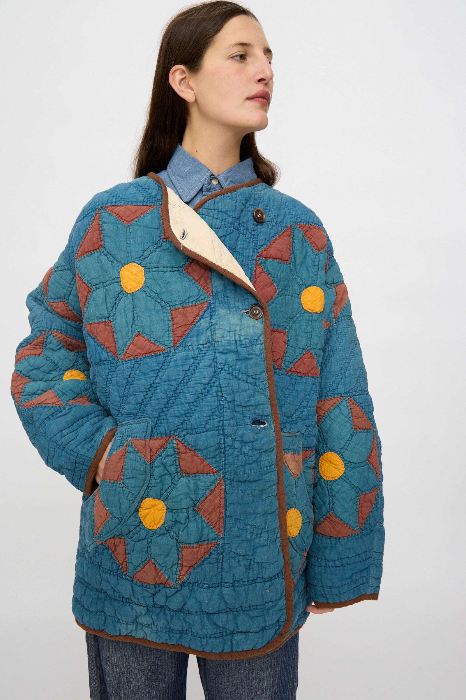 A person wearing the As Ever Vintage Quilt Coat in indigo, brown, and yellow over a denim shirt and blue pants stands against a plain light background.