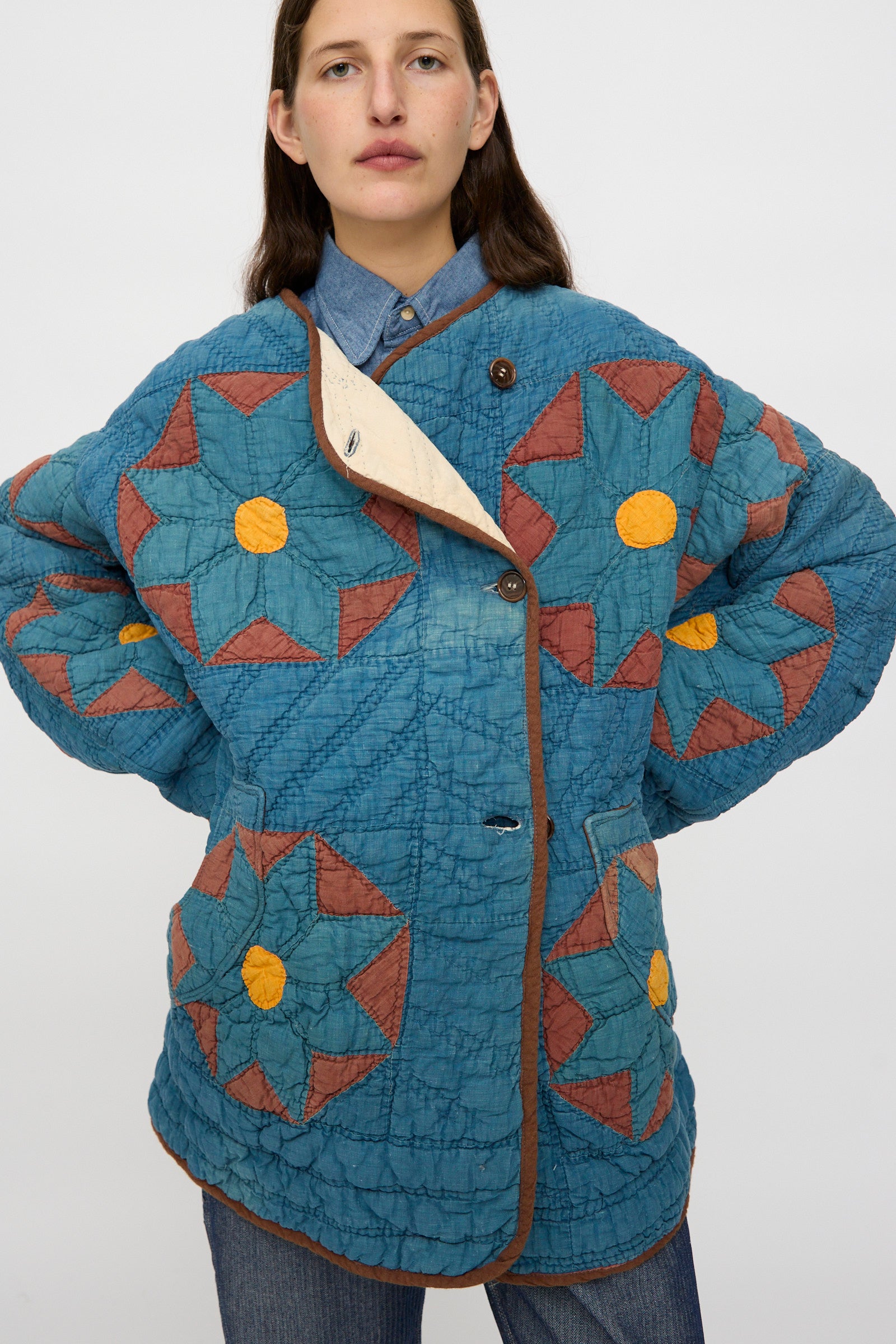 A person wears the As Ever Vintage Quilt Coat in indigo, brown, and yellow over a denim shirt and jeans, standing against a plain background.