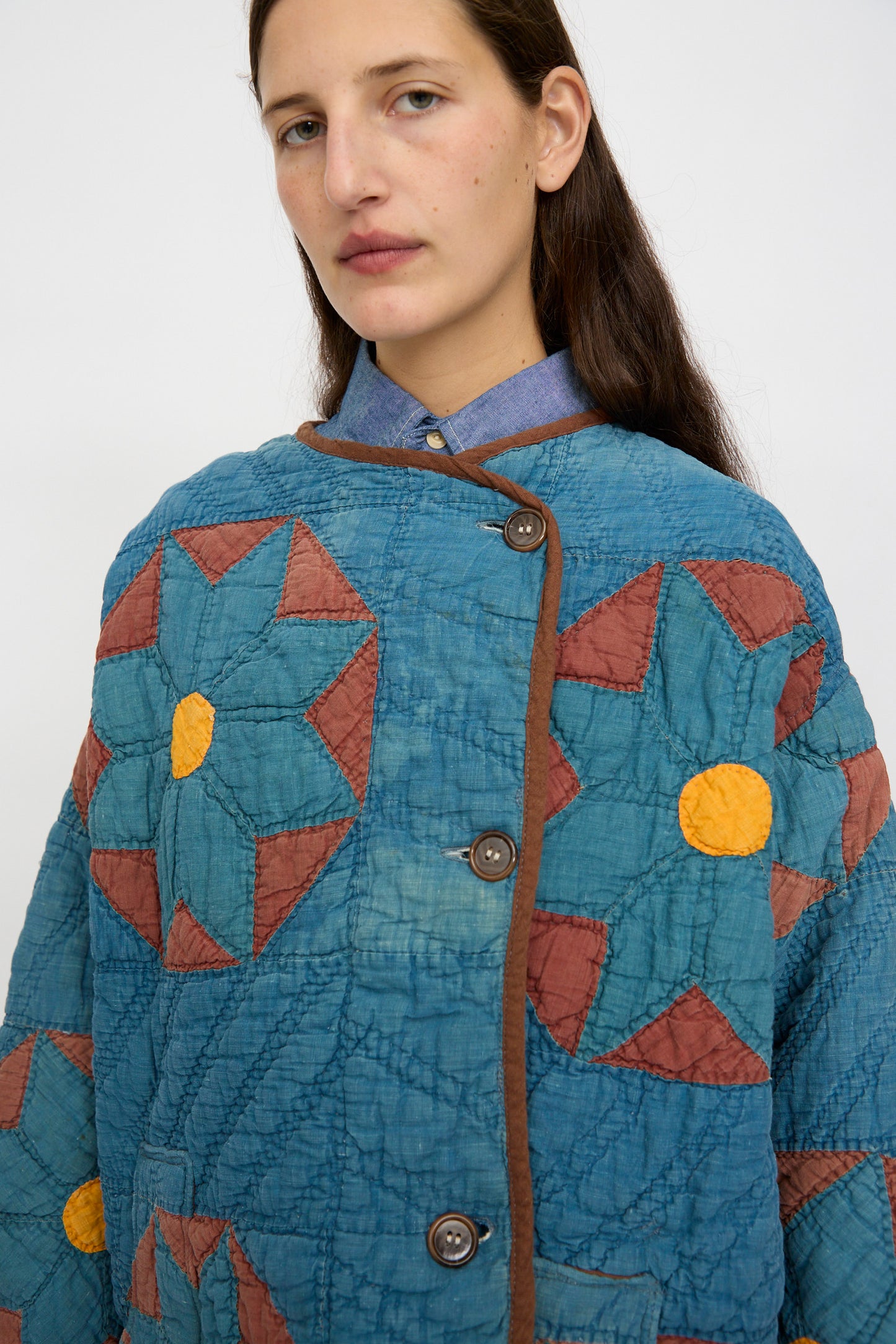 A person wears the As Ever Vintage Quilt Coat in indigo, brown, and yellow over a blue collared shirt.