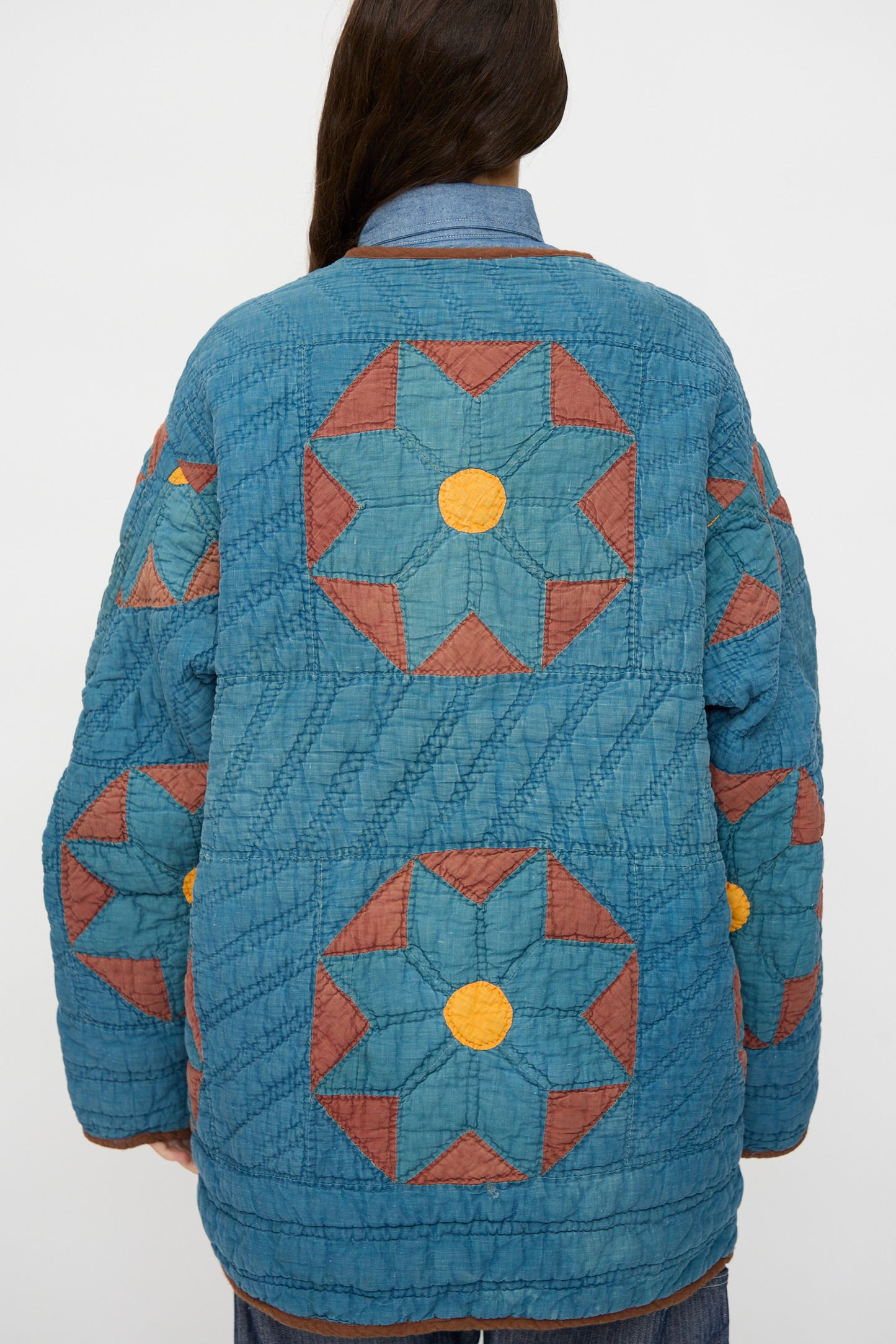 A person faces away from the camera, wearing the As Ever Vintage Quilt Coat in indigo, brown, and yellow with geometric star patterns.