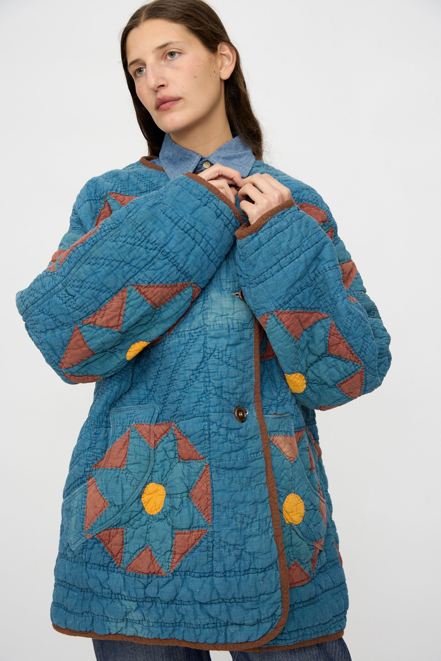 A model wears the As Ever Vintage Quilt Coat in indigo, brown, and yellow with geometric patterns, standing against a plain white background.