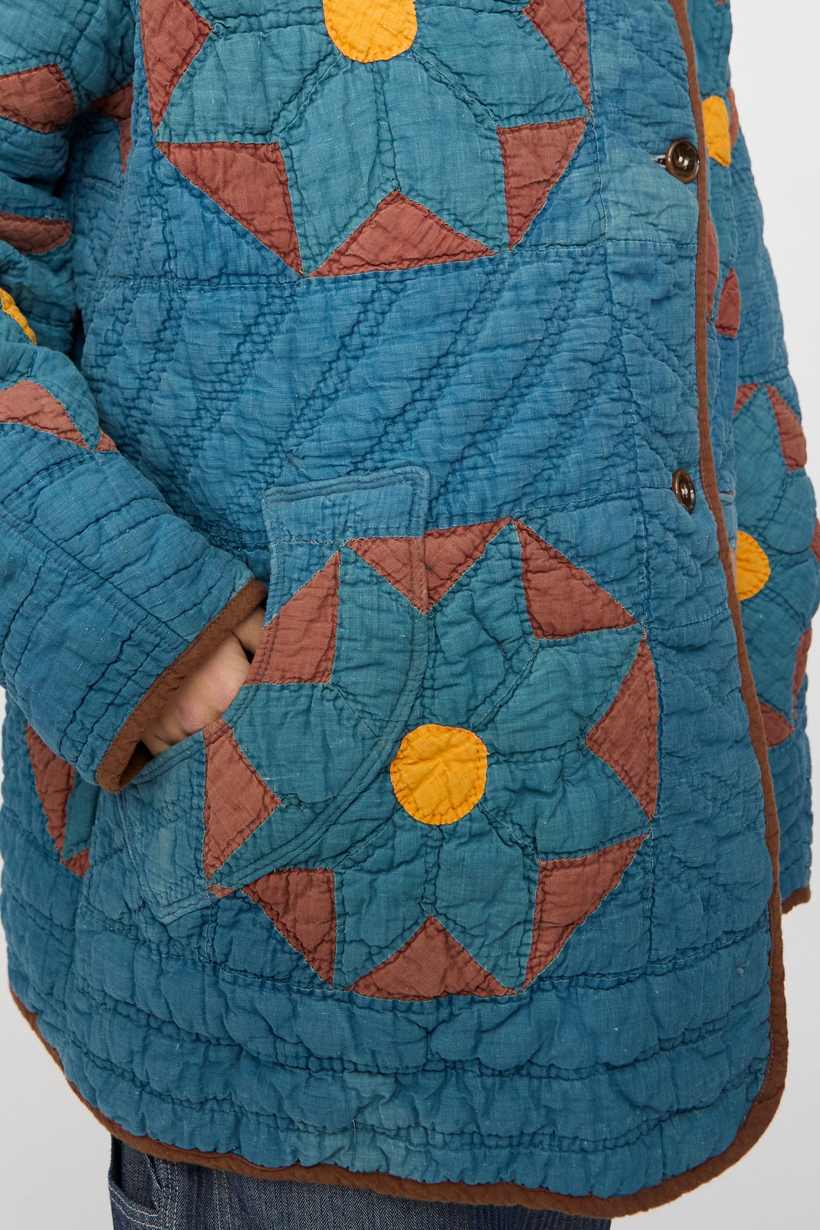 Close-up of a person wearing the As Ever Vintage Quilt Coat in indigo, brown, and yellow, featuring geometric star patterns, with a hand tucked into the pocket.