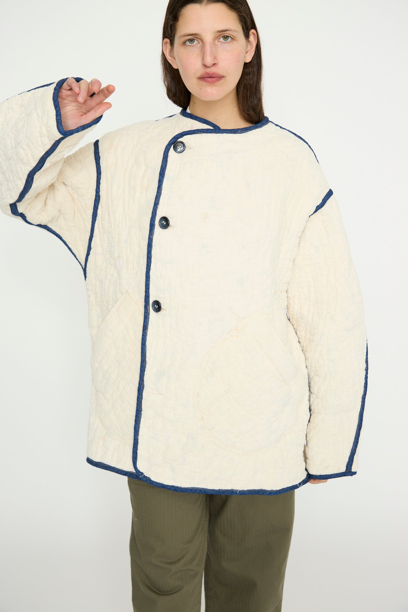 A person models the As Ever Vintage Quilt Coat in Multi Color, an oversized cream jacket with blue trim and dark pants against a plain background. 