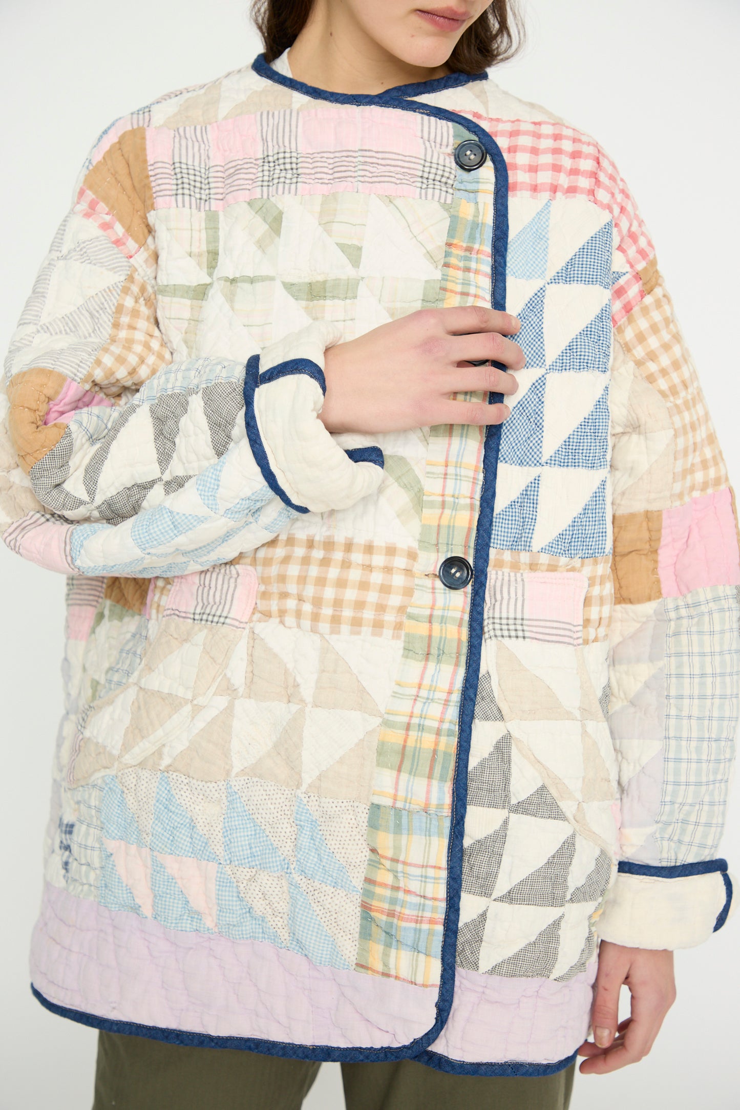 A person wears the Vintage Quilt Coat in Multi Color by As Ever, featuring multicolored geometric patchwork with blue, pink, beige plaid patterns, dark edging, and buttons.