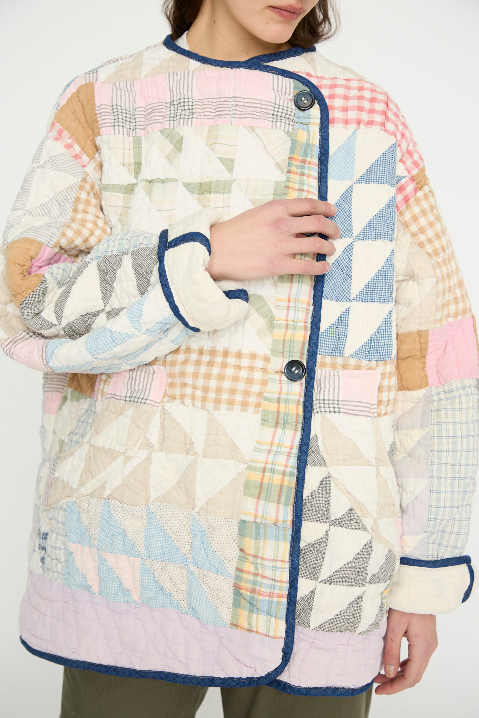 A person wears the Vintage Quilt Coat in Multi Color by As Ever, featuring multicolored geometric patchwork with blue, pink, beige plaid patterns, dark edging, and buttons.