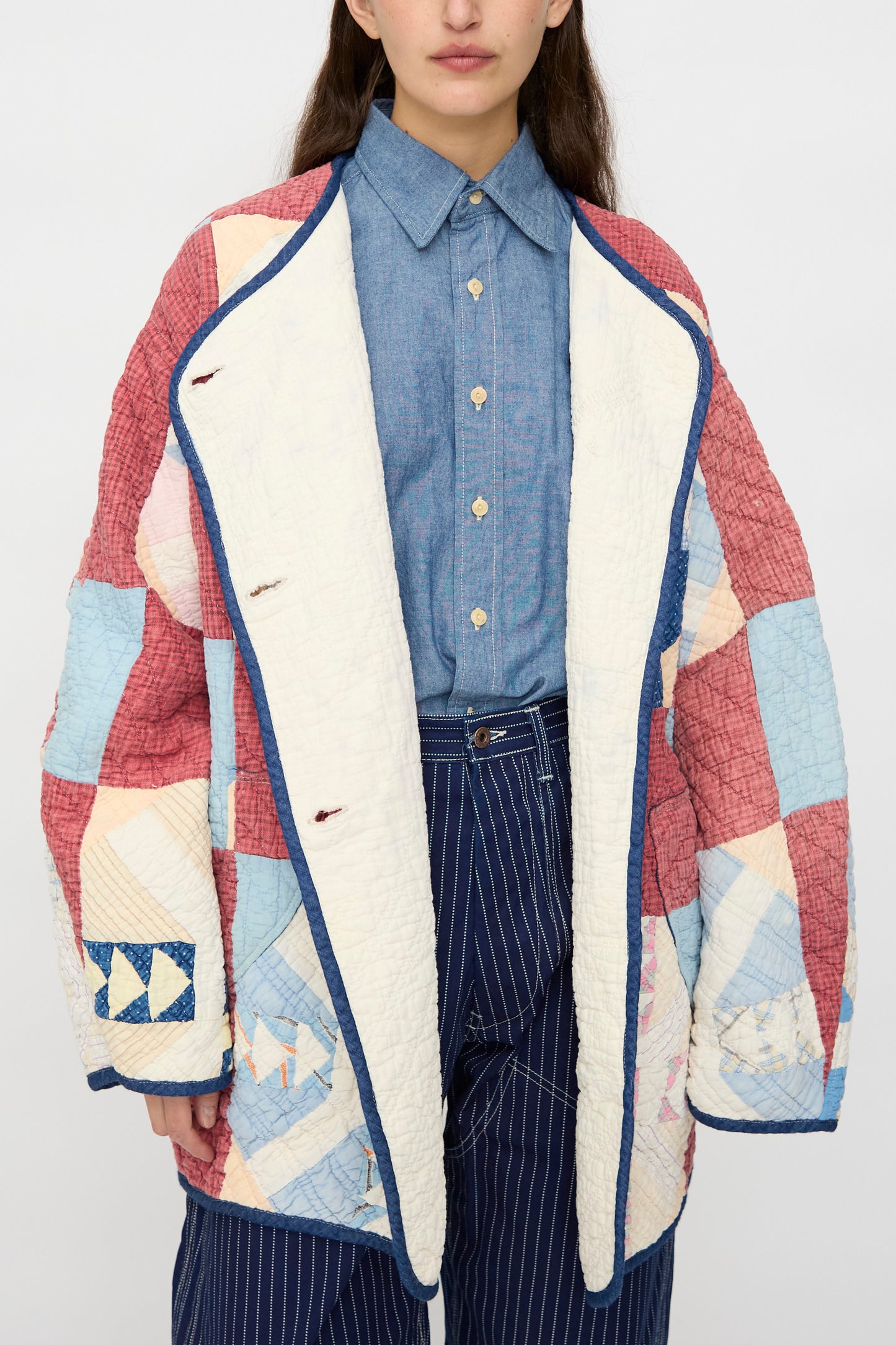 A person models the As Ever Vintage Quilt Coat in Red and Blue, layered over a blue button-up shirt and pinstriped pants, shown from mid-face to thigh against a plain background.