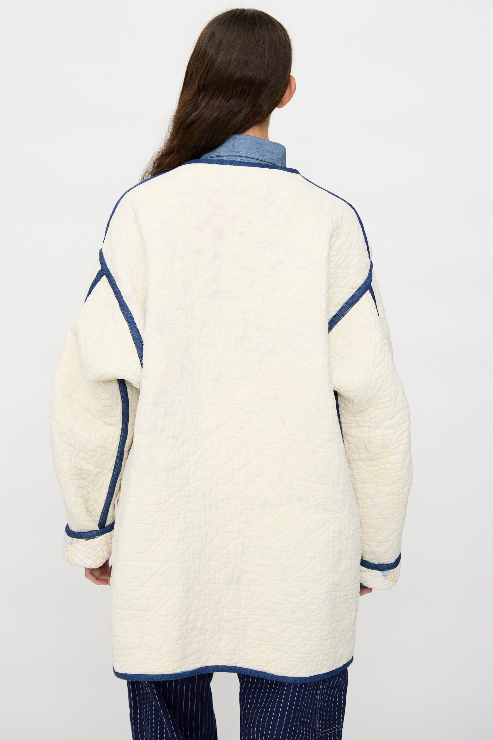 A person with long brown hair stands with their back to the camera wearing As Ever’s Vintage Quilt Coat in Red and Blue, paired with a blue collared shirt and pinstriped pants.