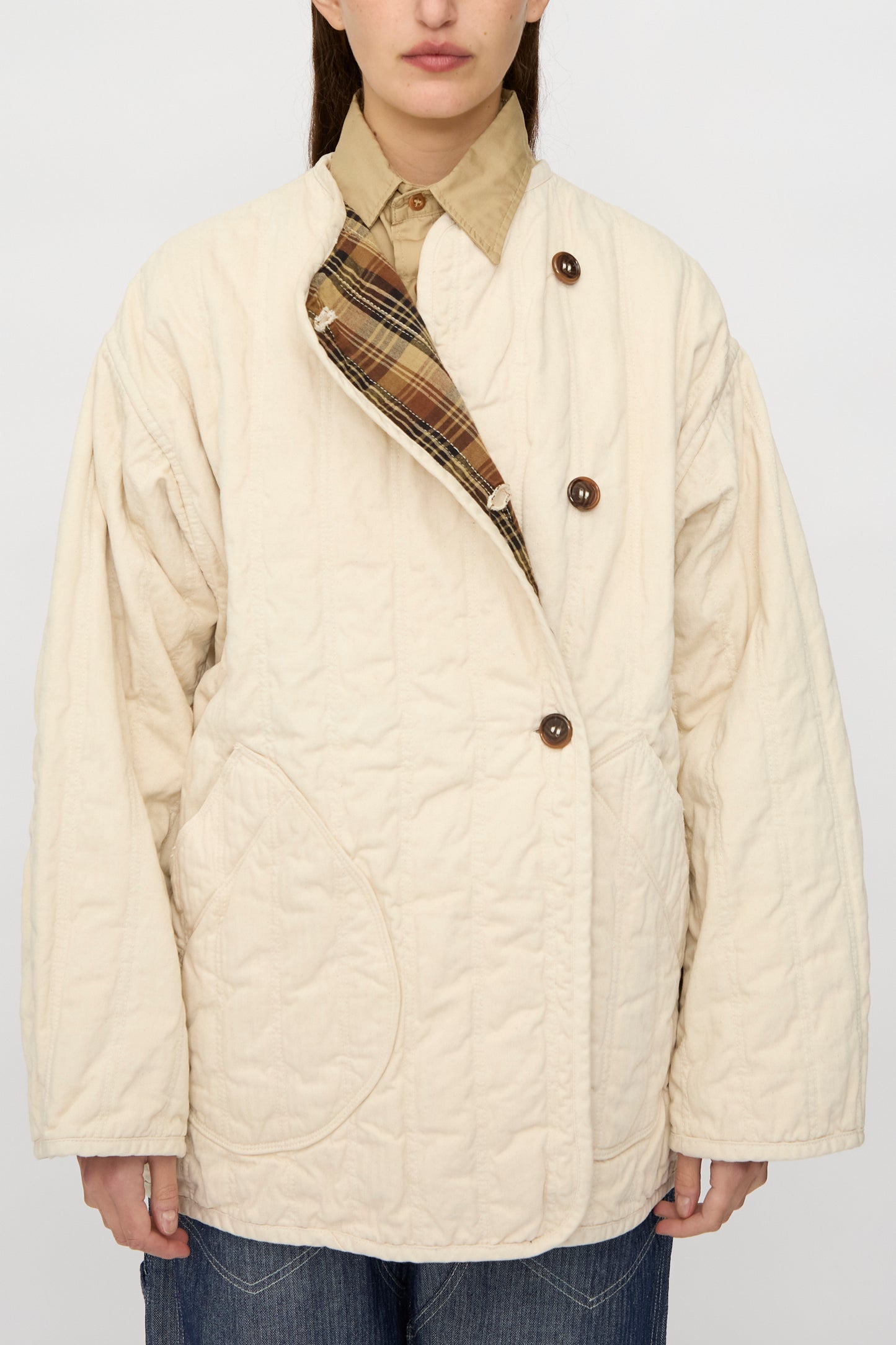 A person wears the As Ever Quilt Coat in HBT Natural—an oversized cream quilted jacket with cotton batting, brown buttons, and visible inner plaid lining—over a tan collared shirt and blue jeans.