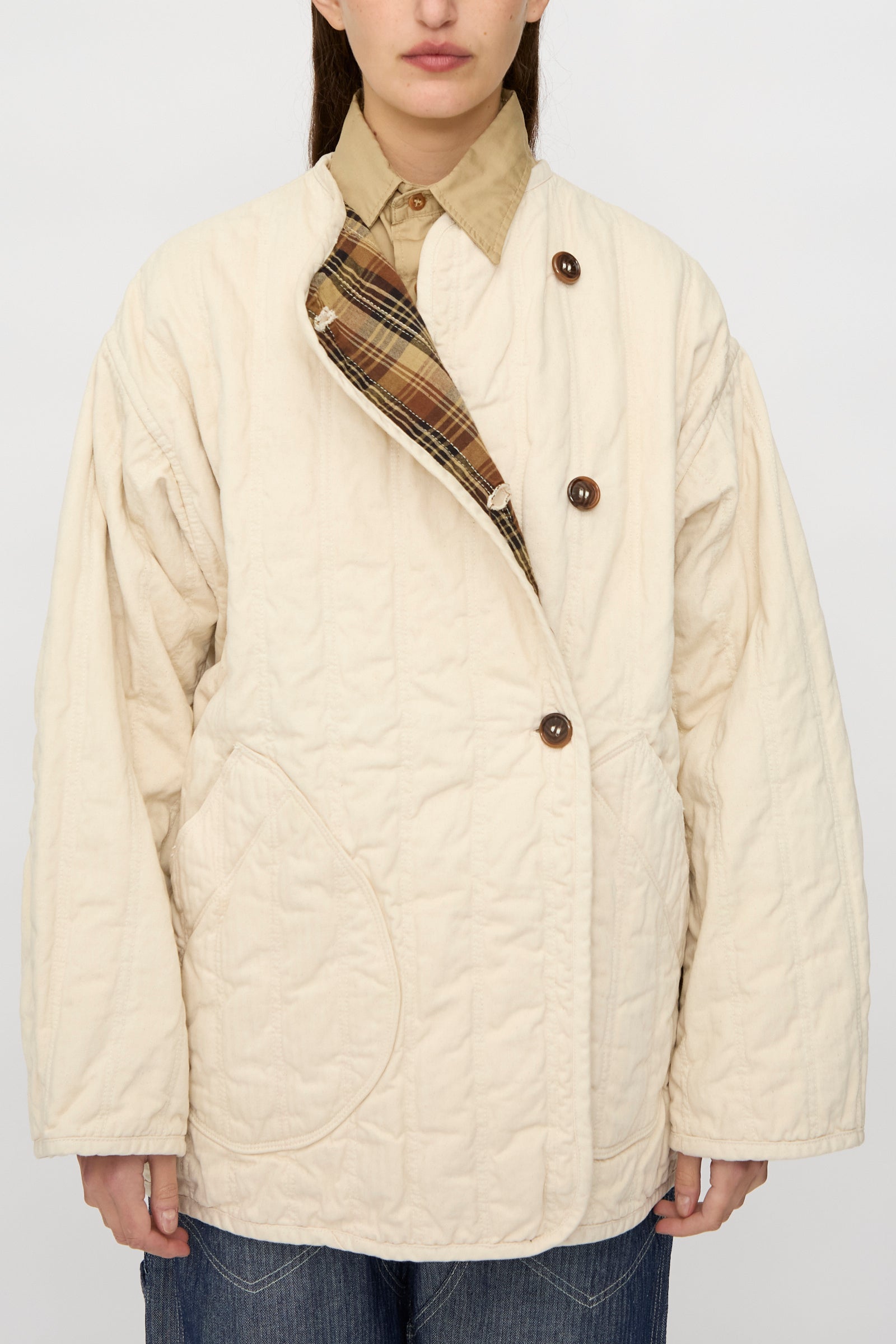 A person wears the As Ever Quilt Coat in HBT Natural—an oversized cream quilted jacket with cotton batting, brown buttons, and visible inner plaid lining—over a tan collared shirt and blue jeans.