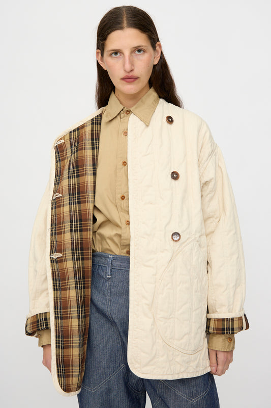 A person wears a tan shirt, blue jeans, and the As Ever Quilt Coat in HBT Natural—an oversized cream quilted jacket with cotton batting and plaid lining, worn open on one side to show off the inner detail.