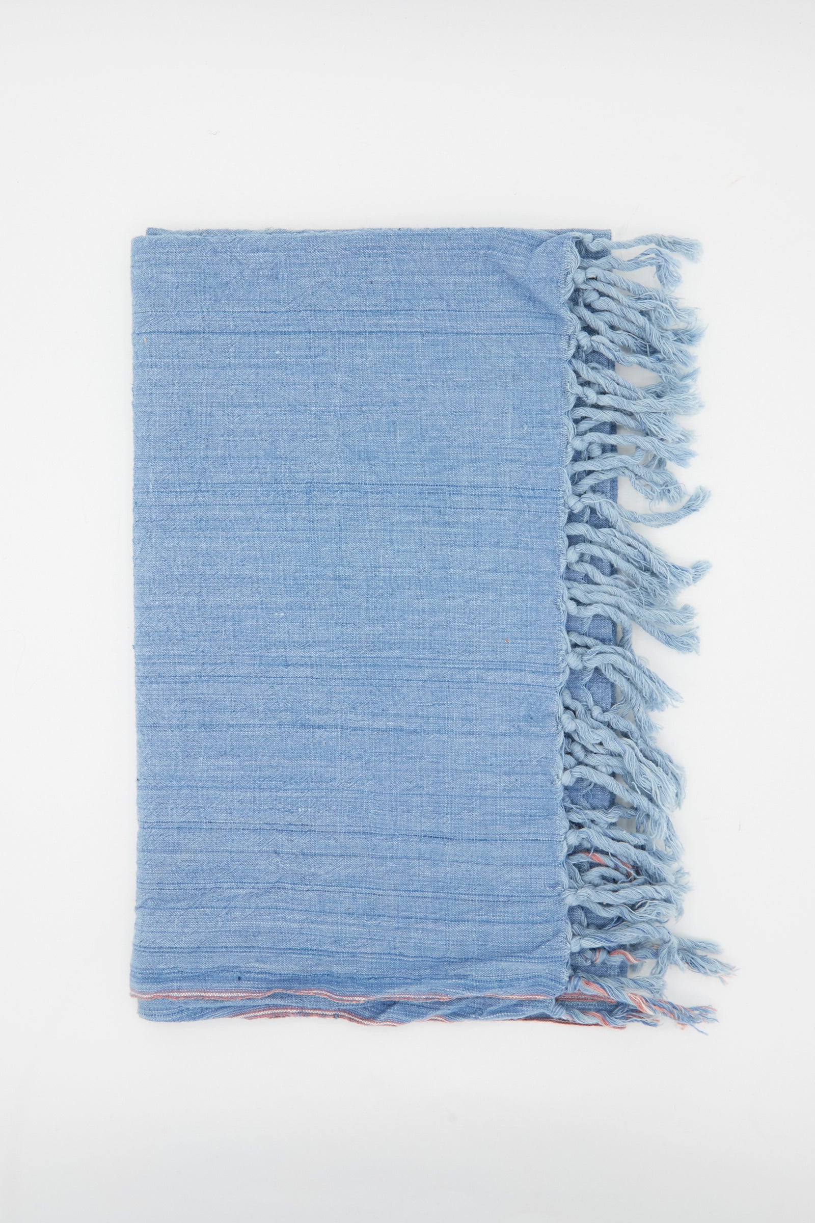 The Auntie Oti Cotton Khadi Red Selvedge Indigo Towel, handwoven in light blue with fringed edges, is shown folded on a white background.