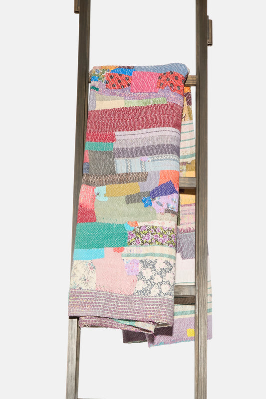 The One of a Kind Vintage Kantha Quilt I by Auntie Oti, made from upcycled cotton, is folded and draped over a wooden ladder set against a plain white background.