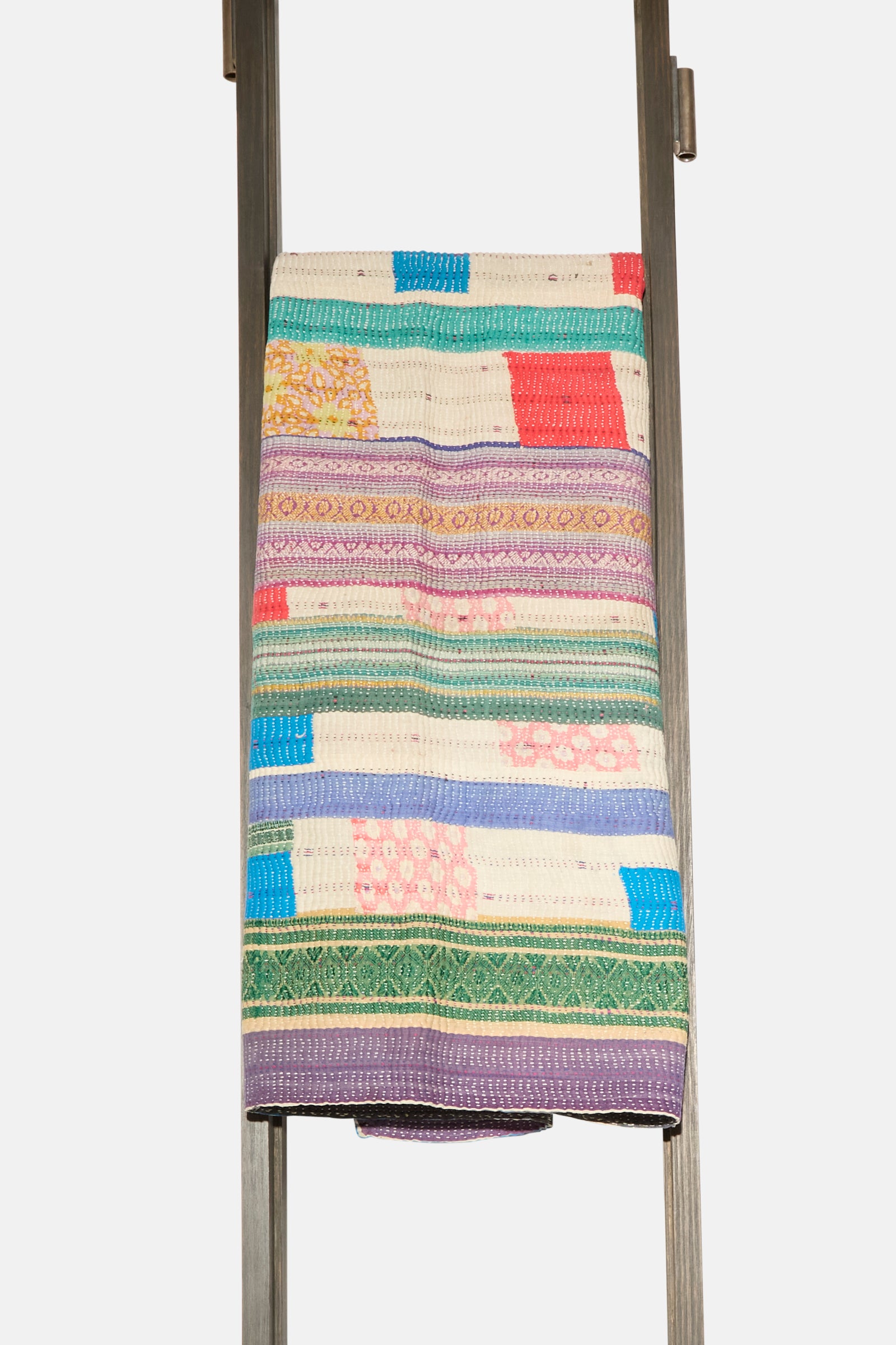 The One of a Kind Vintage Kantha Quilt II by Auntie Oti, made from upcycled cotton with assorted patterns and stripes, is displayed draped over a wooden ladder against a plain background.