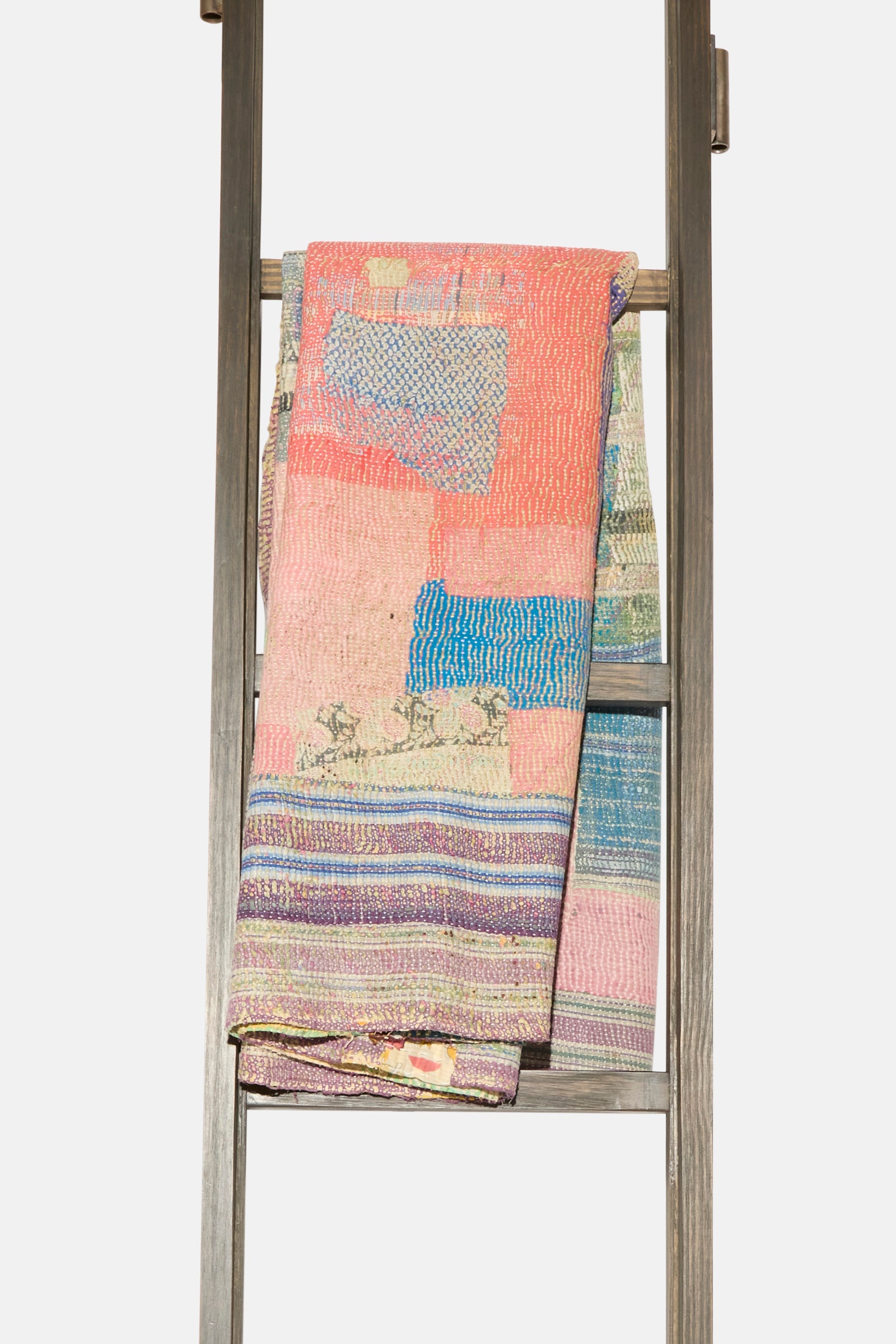 The One of a Kind Vintage Kantha Quilt III by Auntie Oti, featuring upcycled cotton in vibrant patchwork, striped and solid designs, is displayed draped over a wooden ladder against a plain backdrop.
