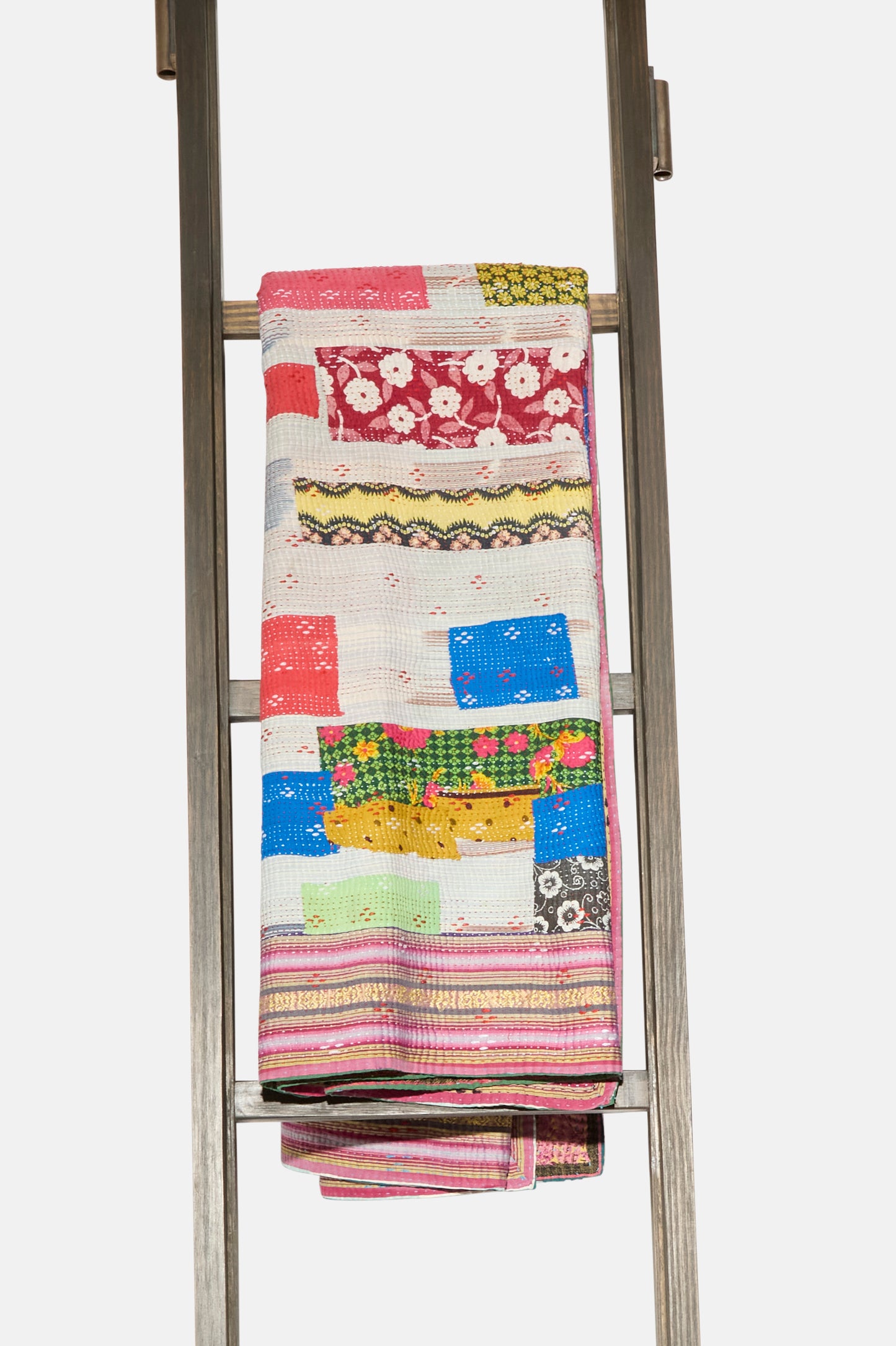 The One of a Kind Vintage Kantha Quilt IV by Auntie Oti, featuring vibrant patterns and colors, is neatly folded and draped over a wooden ladder against a white background.