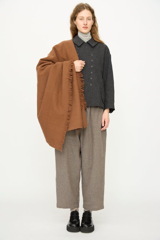 A person stands facing forward, wearing wide-legged gray pants, a dark gray buttoned jacket, and black shoes, with the Auntie Oti Wool Throw in Brown draped over one shoulder.