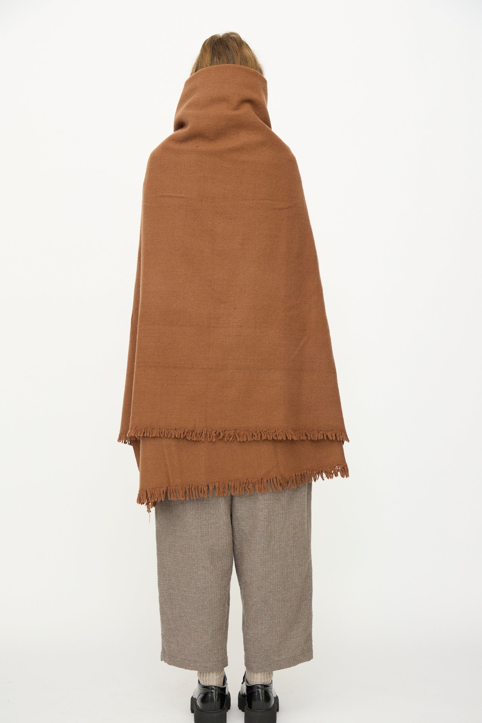 A person stands with their back to the camera, draped in the Auntie Oti Wool Throw in Brown over beige pants, set against a plain white background.