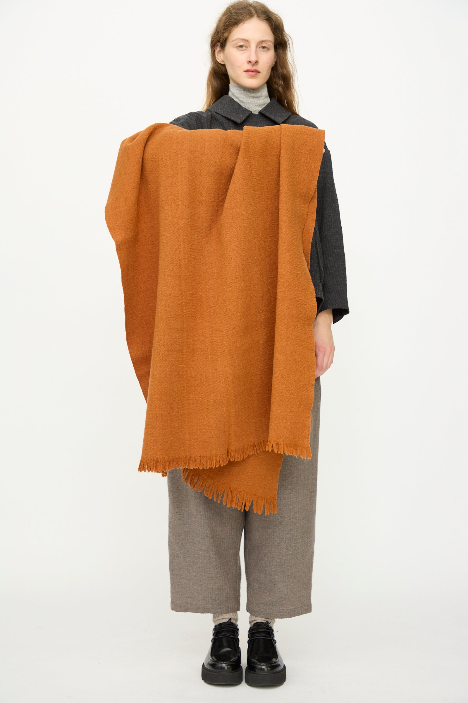 A person stands against a plain background, holding the Auntie Oti Wool Throw in Rust with a fringe edge draped over their shoulder. They wear a grey turtleneck, dark shirt, loose grey pants, and black shoes.
