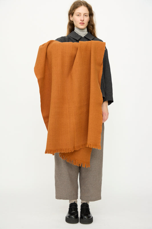 A person stands against a plain background, holding the Auntie Oti Wool Throw in Rust with a fringe edge draped over their shoulder. They wear a grey turtleneck, dark shirt, loose grey pants, and black shoes.