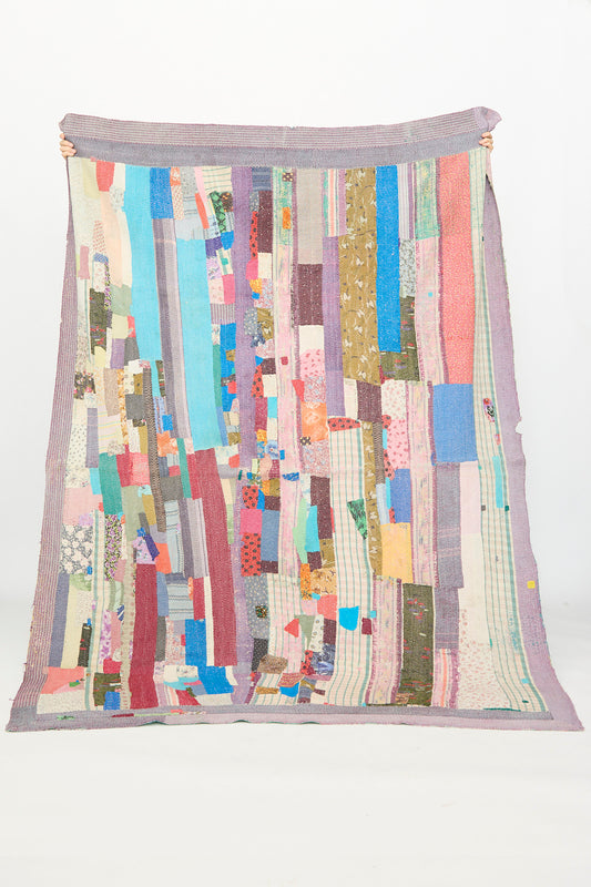 A person holds up the Auntie Oti One of a Kind Vintage Kantha Quilt I, made from upcycled cotton strips arranged in colorful vertical and diagonal patterns on a white background.