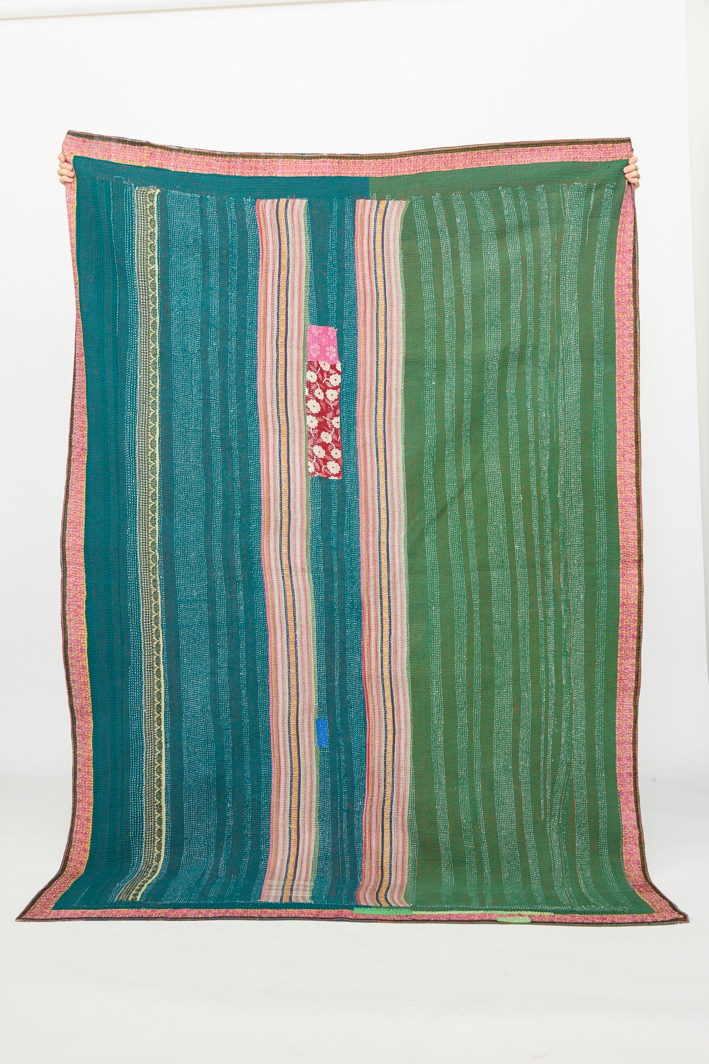 The One of a Kind Vintage Kantha Quilt IV by Auntie Oti features vertical green, blue, and pink stripes, a central pink floral patch, and a pink border. It’s displayed upright against a plain white background.