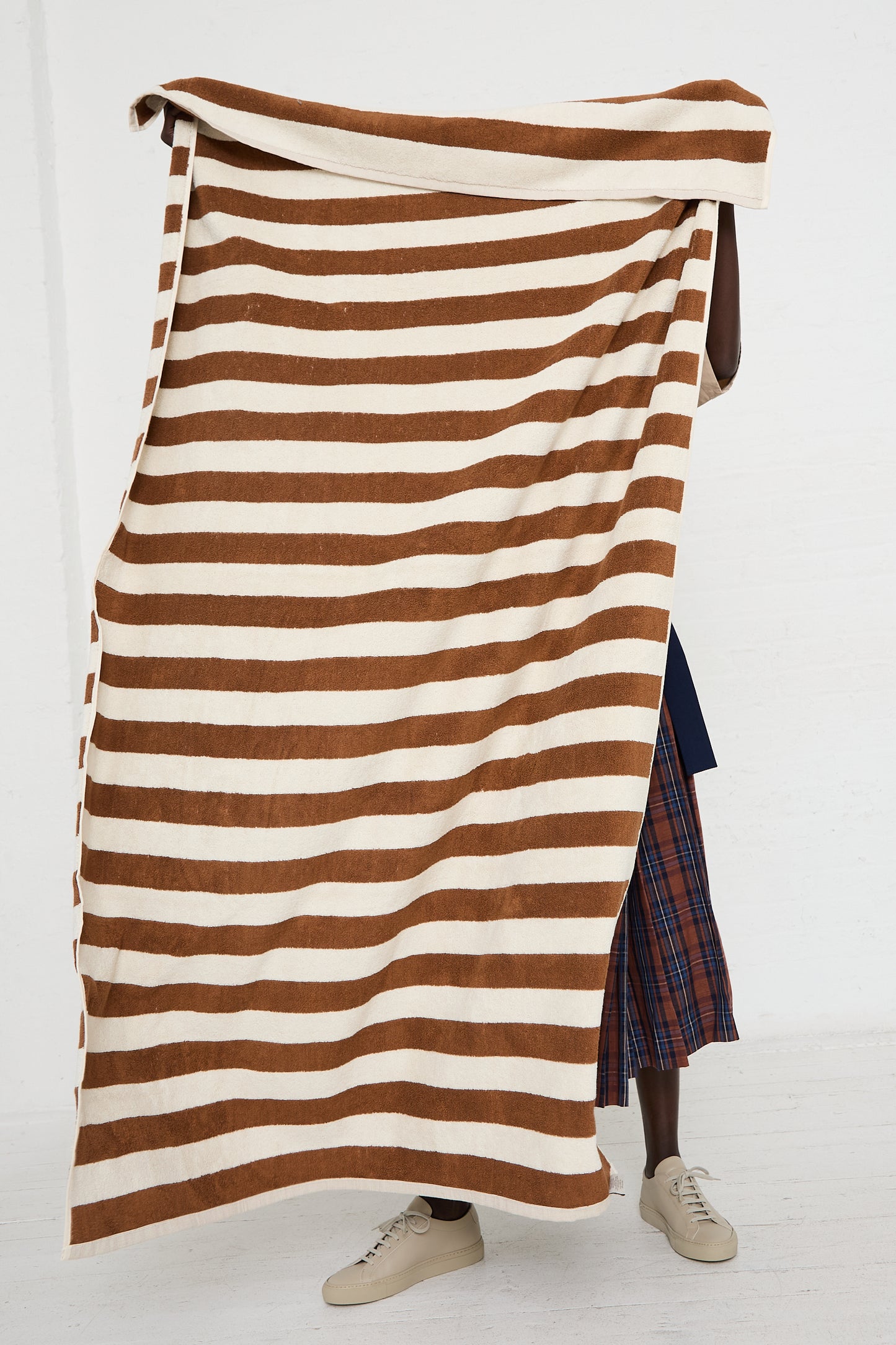 Person holding up a large Autumn Sonata Maria Pool Towel with wide brown and white stripes, reminiscent of traditional carpet making, partially covering their body, against a plain background.