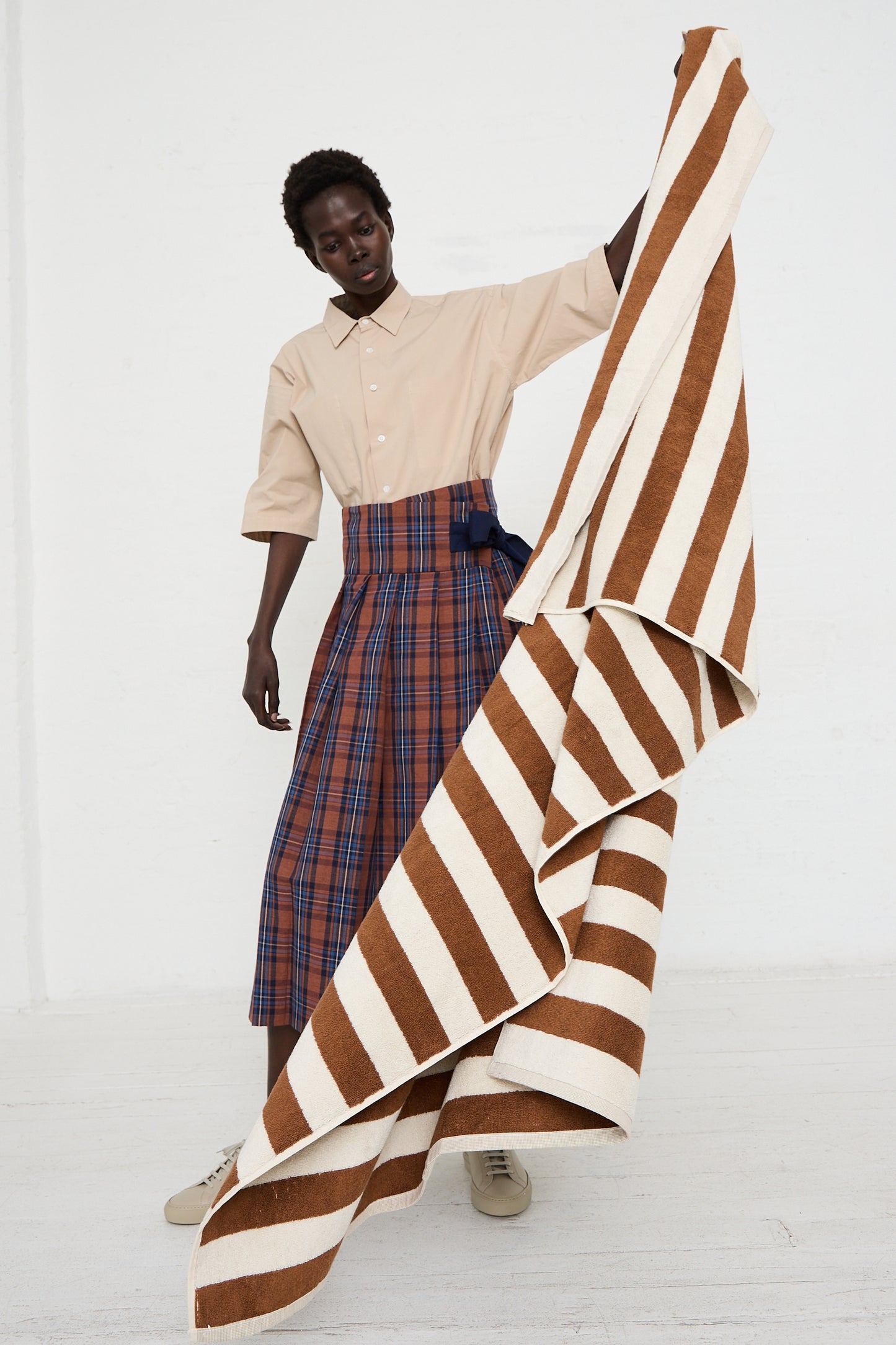 A person stands indoors holding an oversized Autumn Sonata Maria Pool Towel made from organic cotton. They wear a beige shirt, a plaid skirt, and beige shoes. The background is plain white.