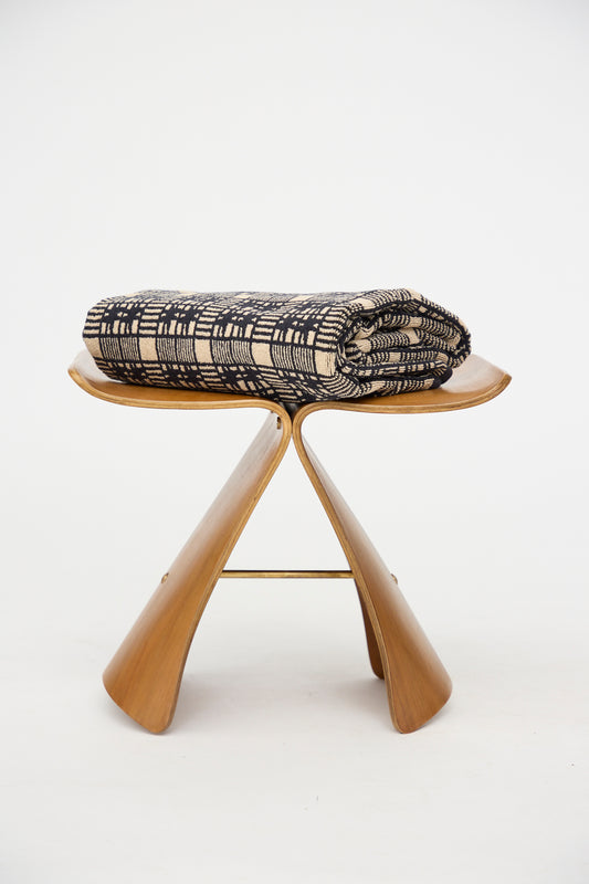 The Alma Bath Sheet by Autumn Sonata, featuring a folded black and beige pattern in organic cotton, rests on a wooden butterfly-style stool against a plain white background.