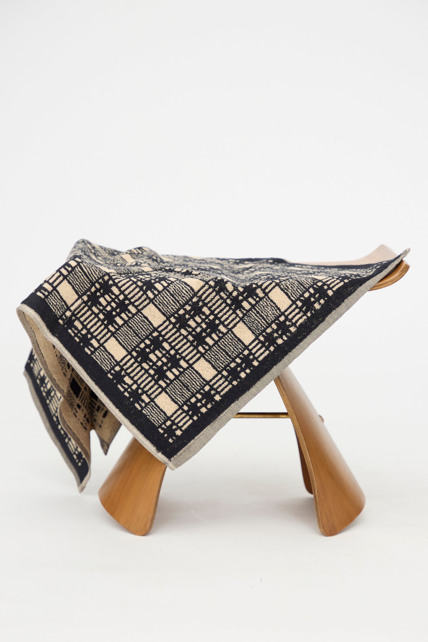 The Alma Hand Towel by Autumn Sonata, crafted from organic cotton and featuring a black and beige geometric pattern, is elegantly draped over a modern wooden stool against a plain white background. 