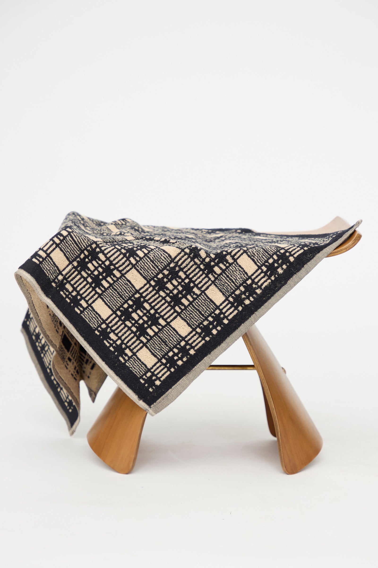 The Alma Hand Towel by Autumn Sonata, crafted from organic cotton and featuring a black and beige geometric pattern, is elegantly draped over a modern wooden stool against a plain white background. 