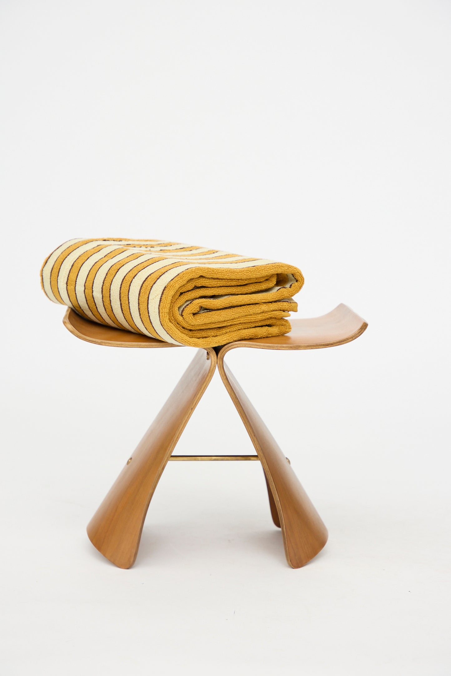The Helena Pool Towel in Ochre by Autumn Sonata, featuring ochre and brown stripes inspired by Edo textiles and crafted from organic cotton, is folded neatly on a wooden butterfly stool against a plain white background.