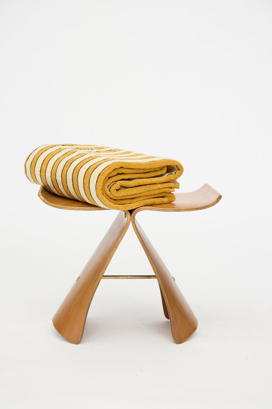 The Helena Pool Towel in Ochre by Autumn Sonata, featuring ochre and brown stripes inspired by Edo textiles and crafted from organic cotton, is folded neatly on a wooden butterfly stool against a plain white background.