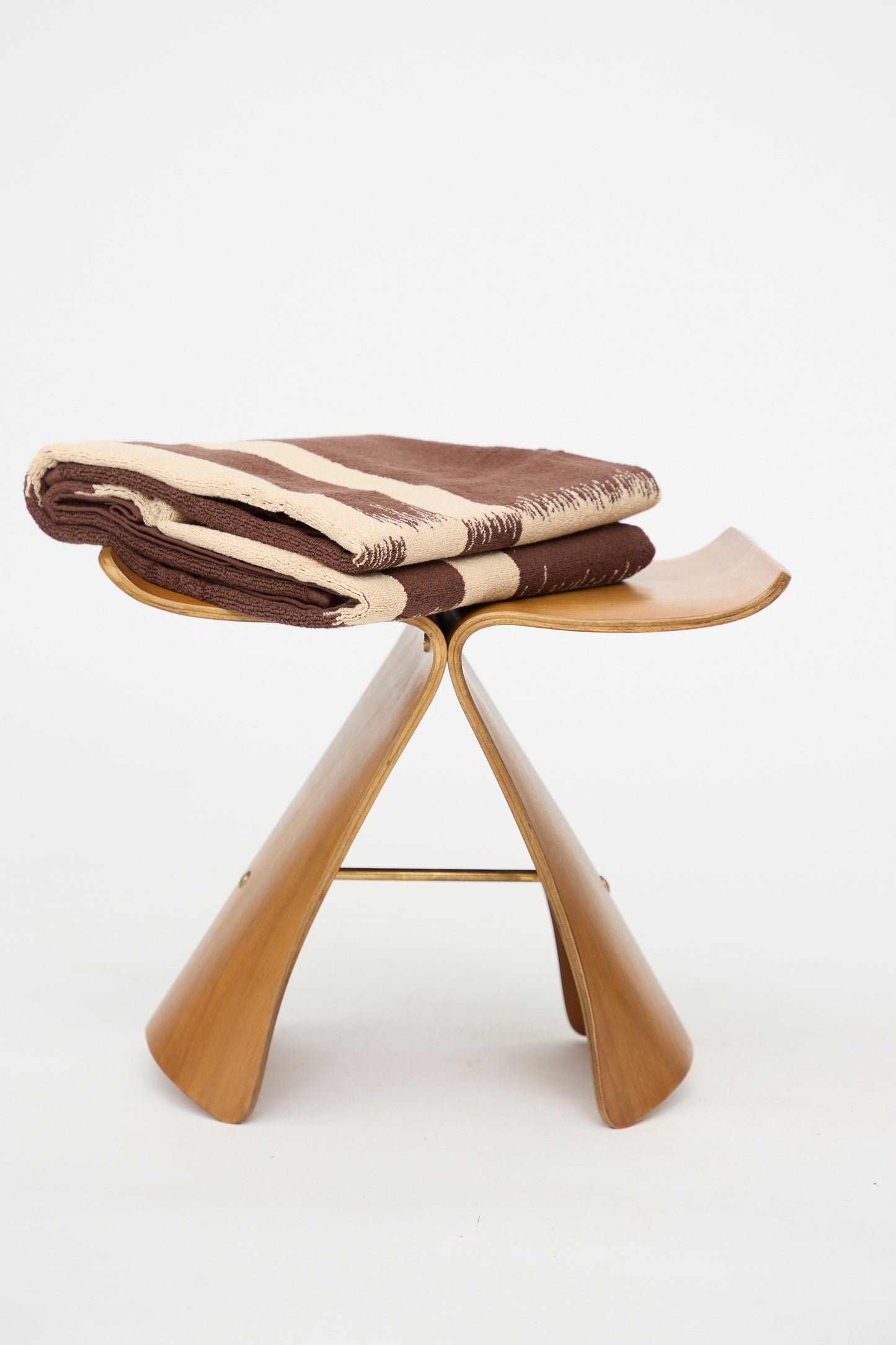 A folded Karin Bath Sheet by Autumn Sonata, featuring brown and beige stripes and crafted from organic cotton using the traditional Katazome technique, rests on a wooden, butterfly-shaped stool against a plain white background.